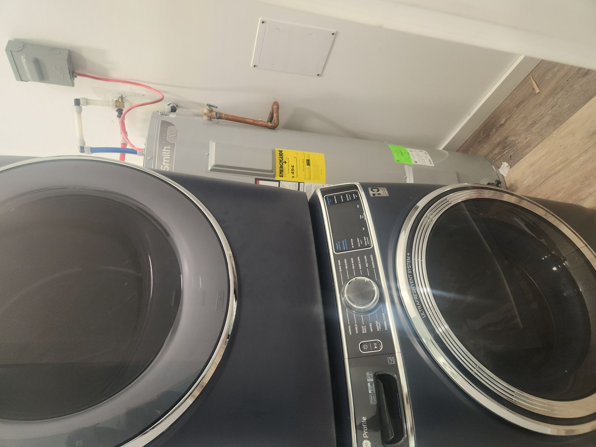 Two front-loading laundry machines under a utility ceiling, with exposed pipes and wiring above.