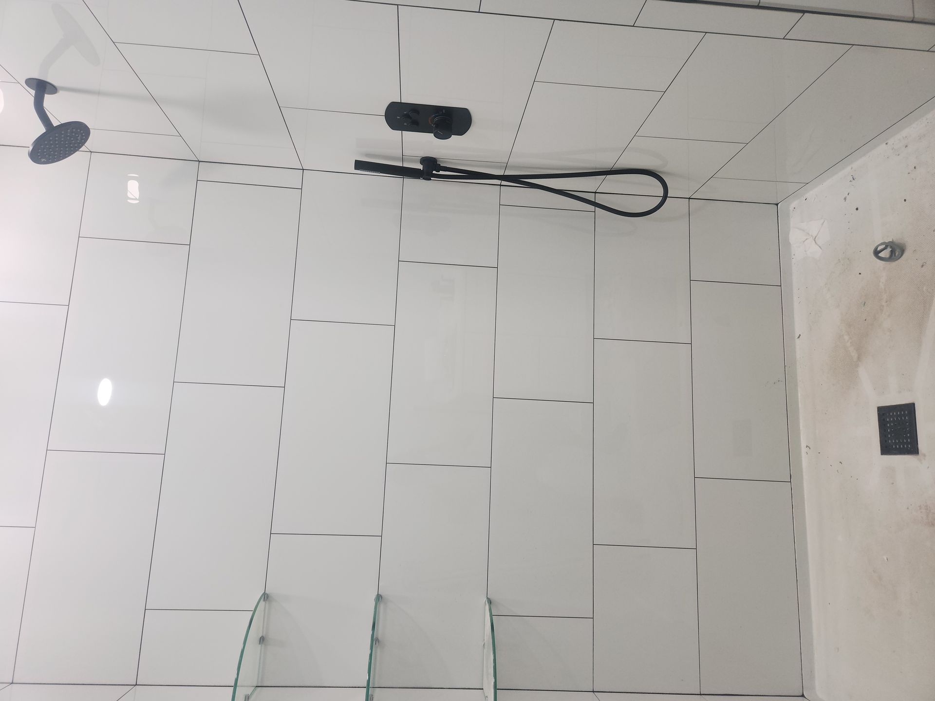 Partially tiled bathroom wall and ceiling with a shower fixture and black hose, under construction
