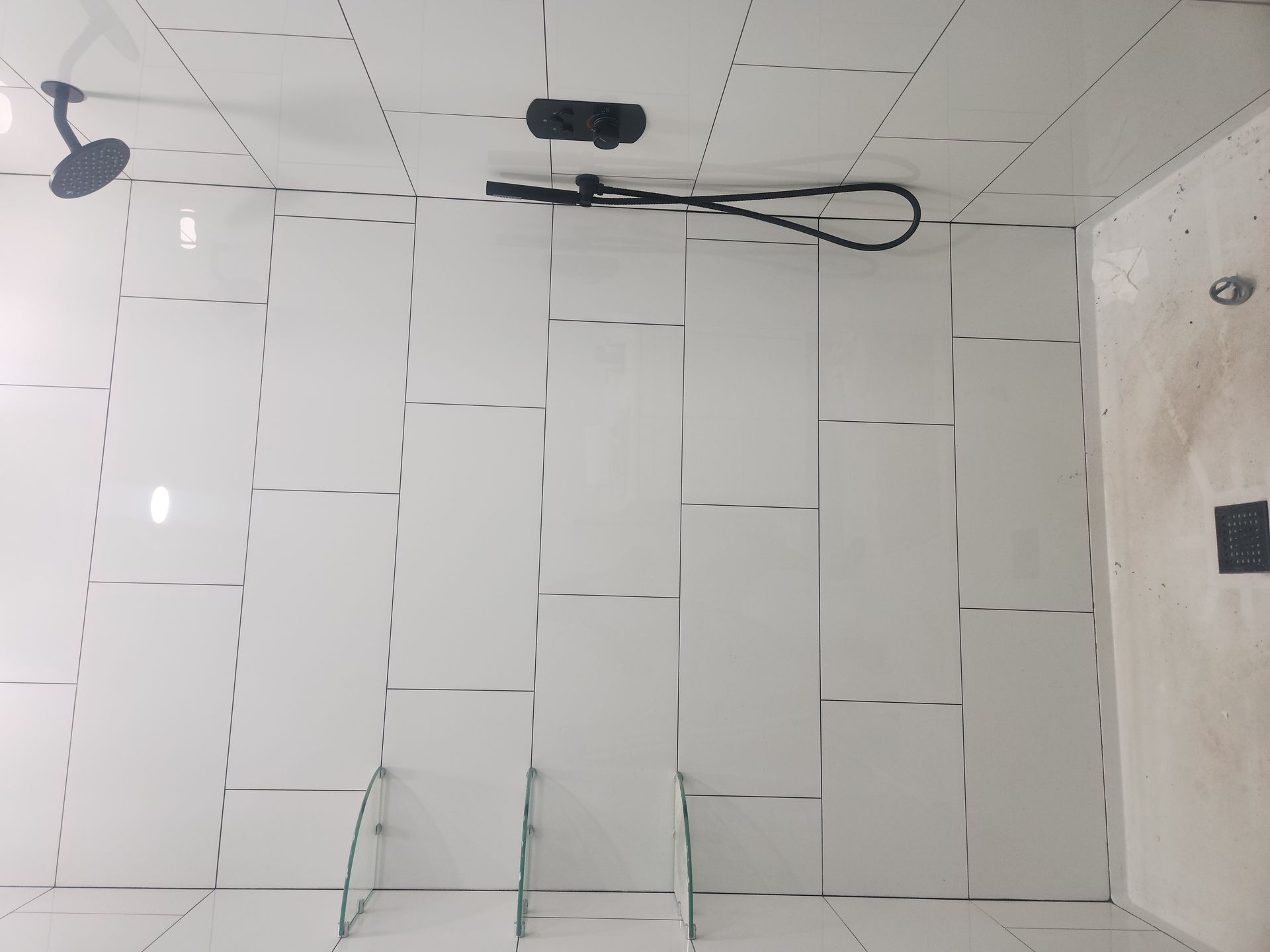 White tiled bathroom wall with a black shower hose and fixture near the ceiling.