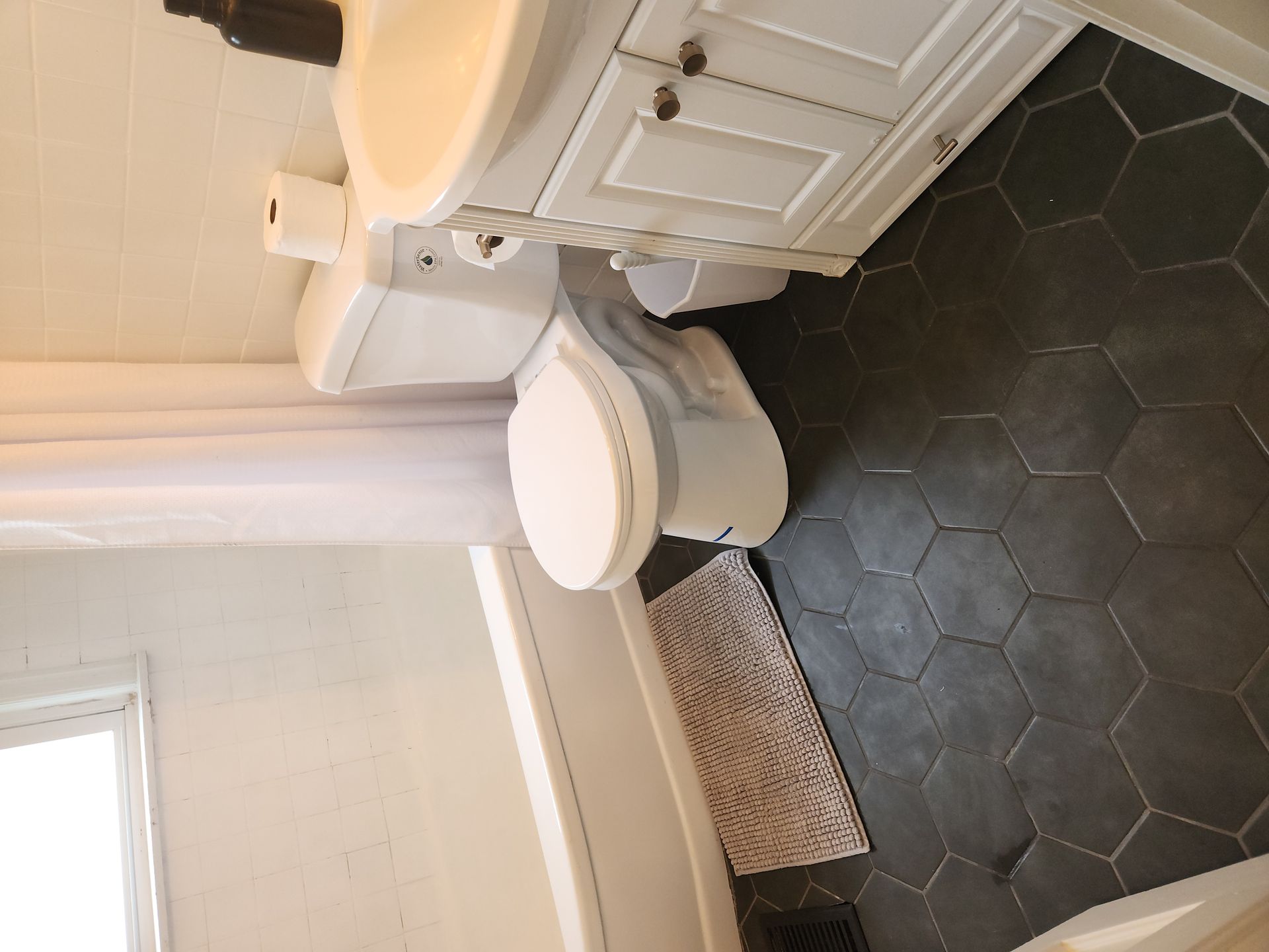 Small bathroom with white vanity, toilet, and black hex tile floor under a window.