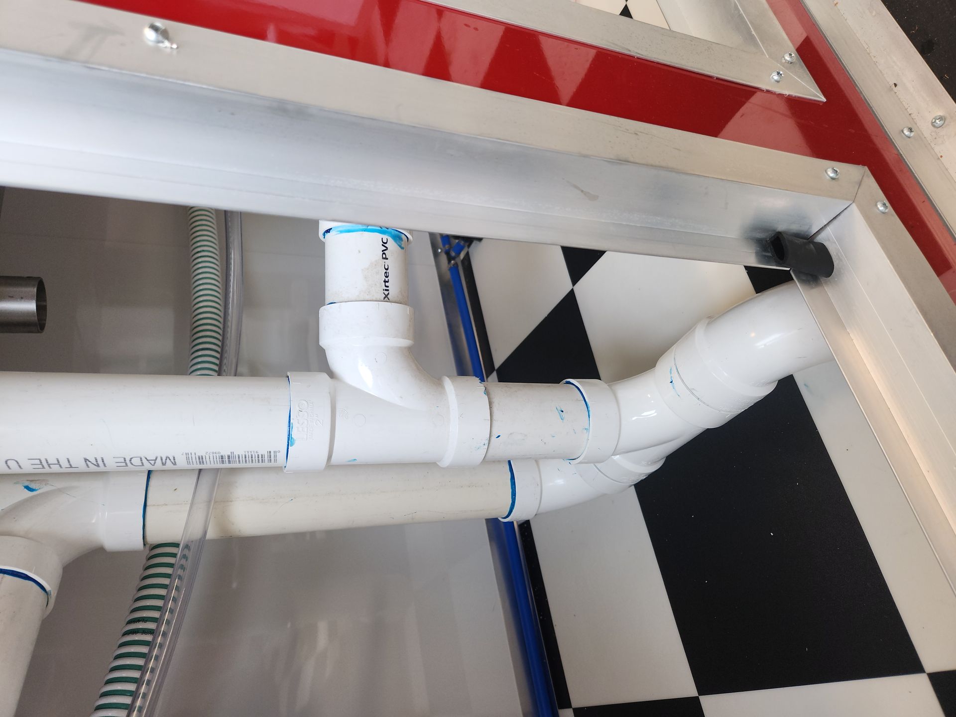 White PVC pipes with elbows and joints under a sink against black-and-white checkered flooring.