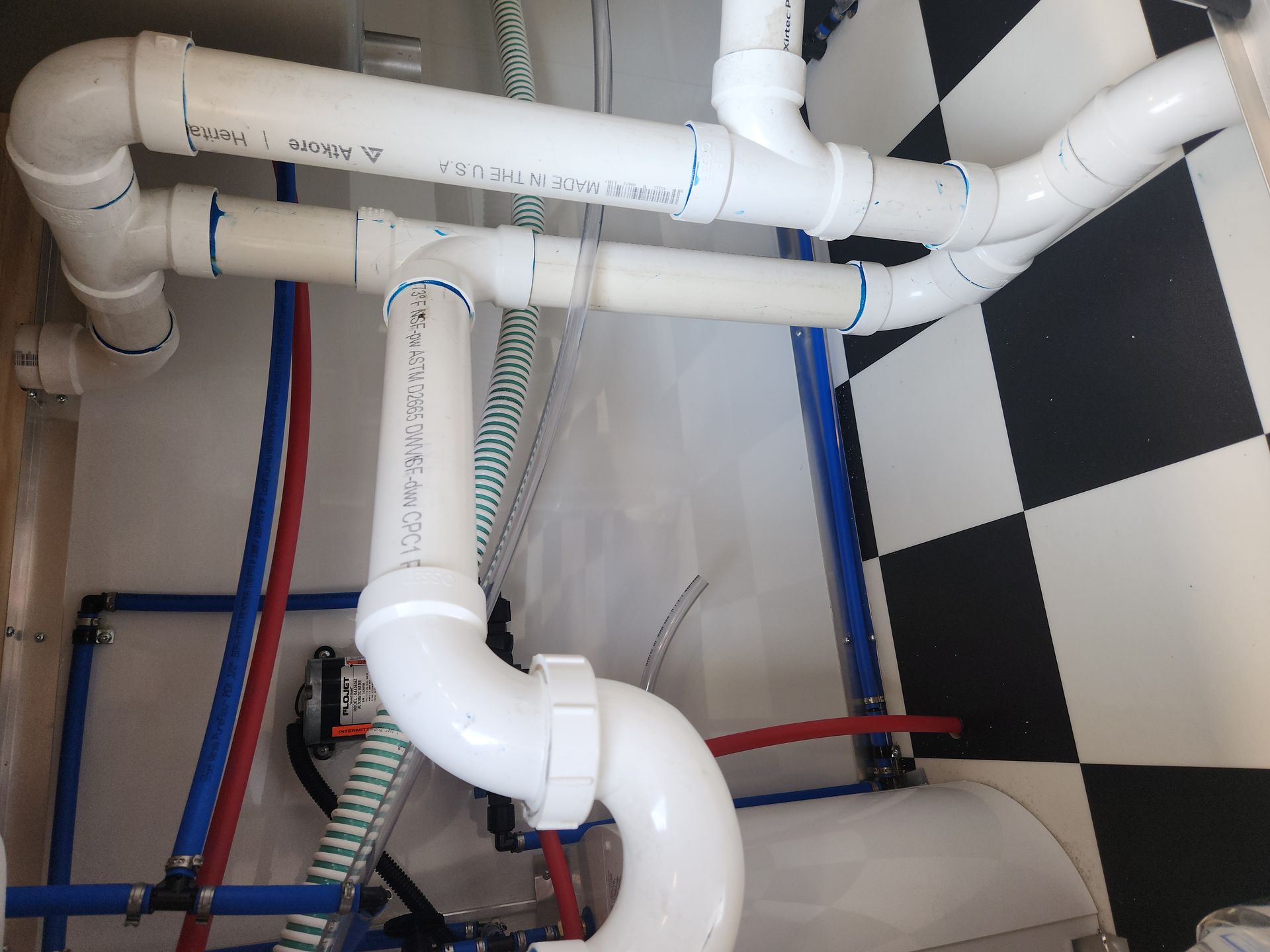 White PVC plumbing pipes and hoses under a sink against a black-and-white checkered wall.