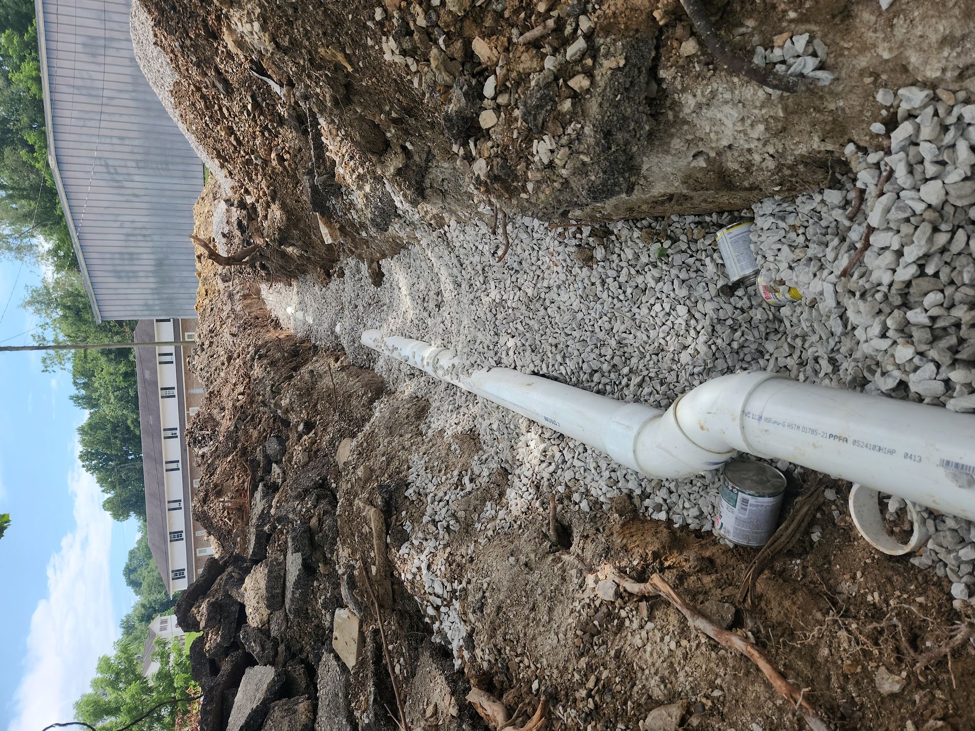 White drainage pipe runs through a dirt trench beside a gravel pile and building foundation.