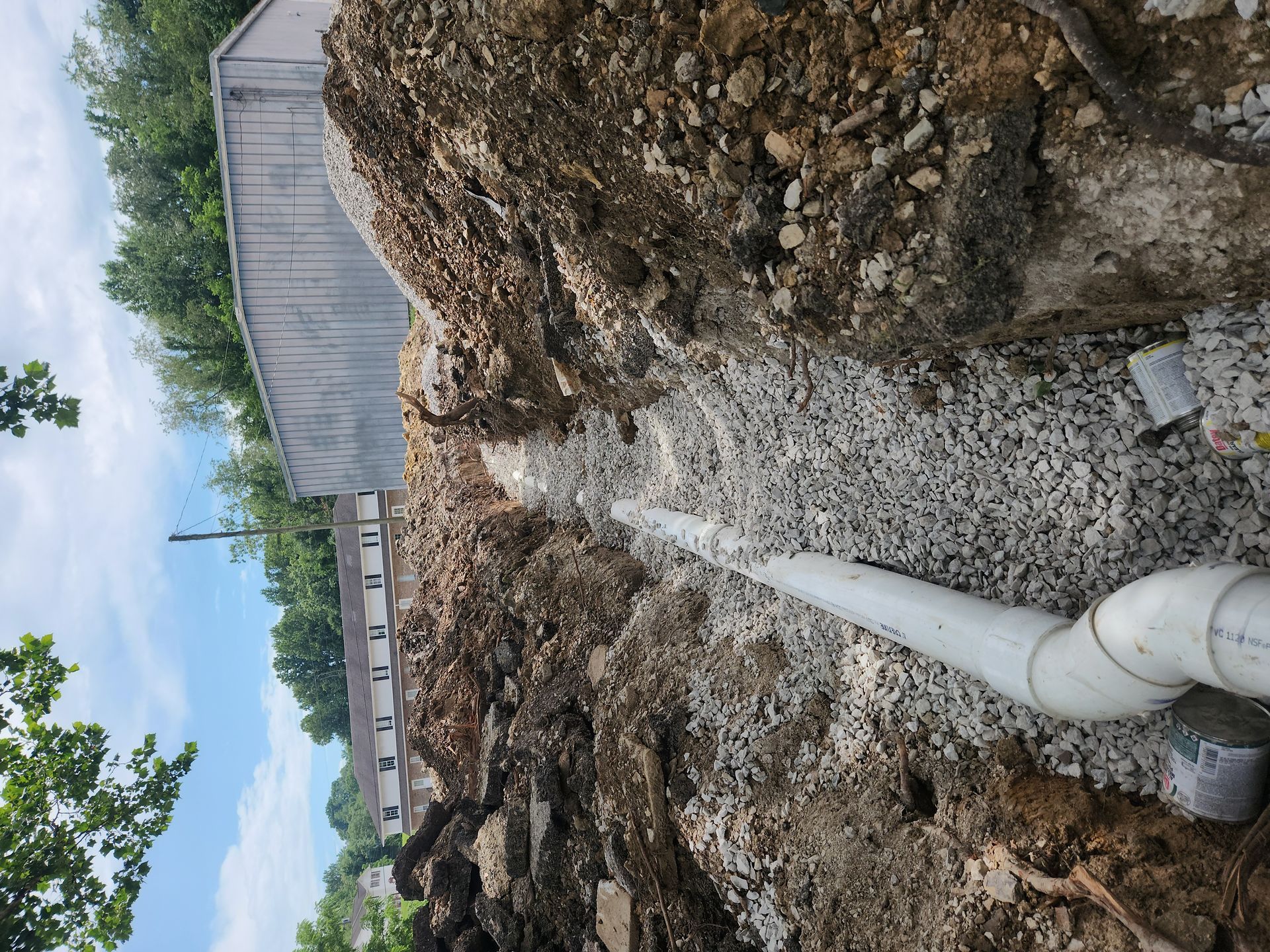 Excavation trench beside a building with a white pipe running through gravel and dirt, under a cloudy sky