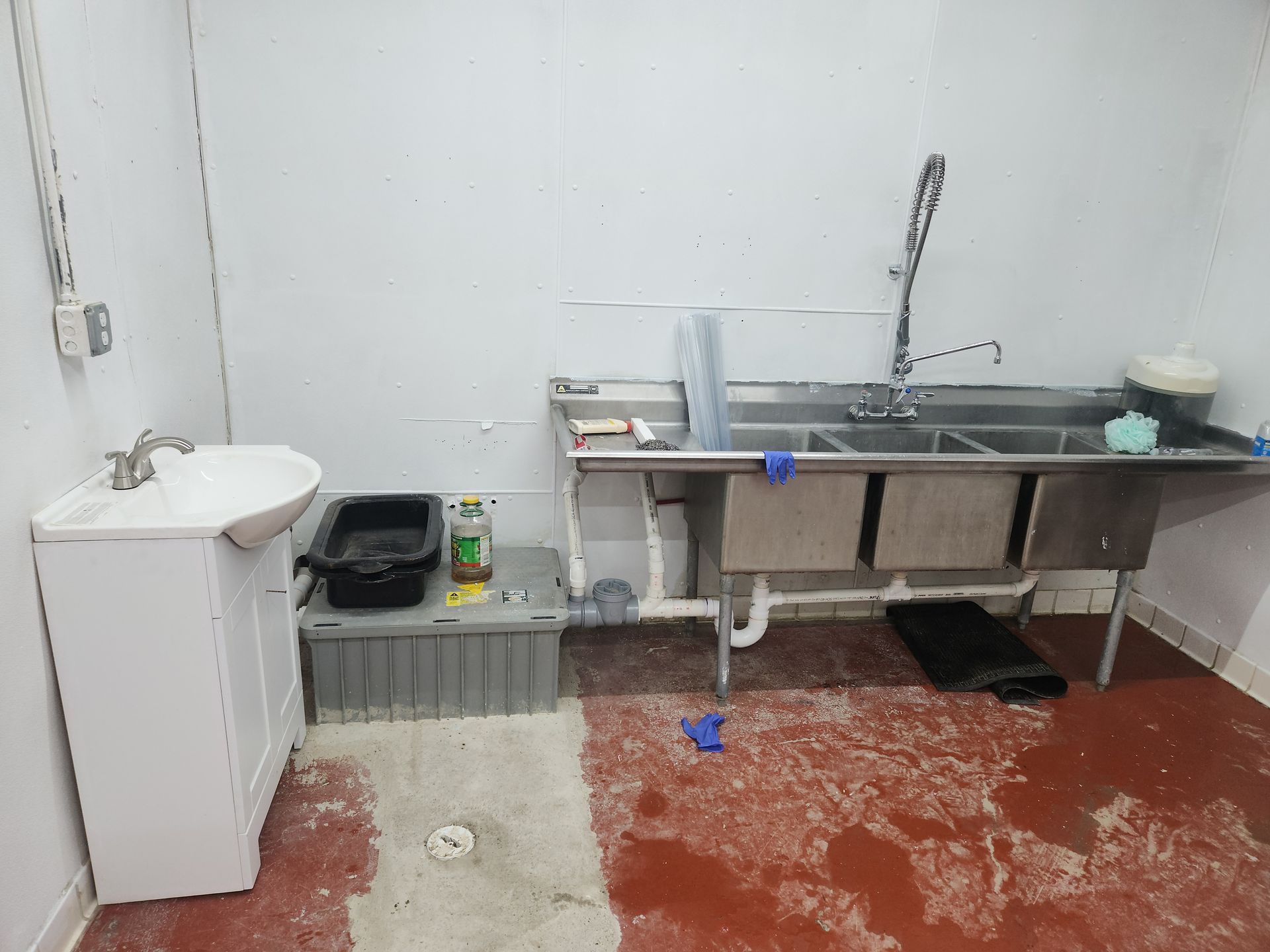 Small utility room with sink, cabinet, red floor, trash bin, and cleaning supplies.