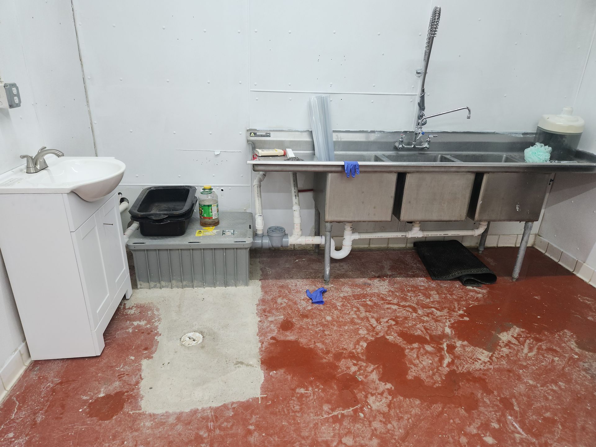A small industrial sink area with a stainless counter, white cabinet, buckets, and a red-speckled floor.