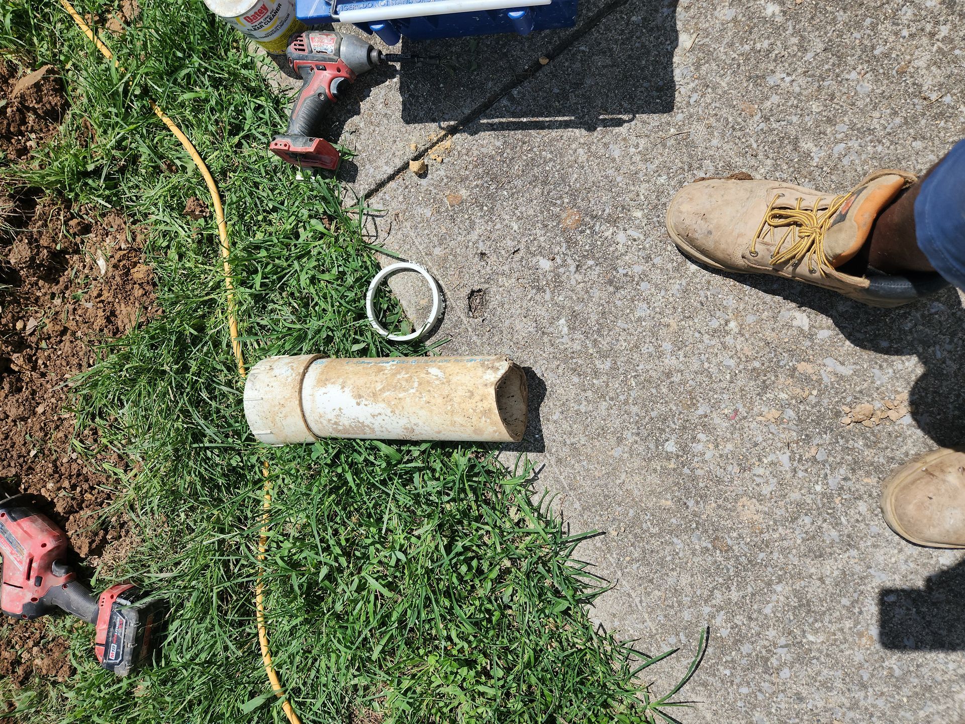 Tools and boots on grass beside a cracked concrete pipe and coiled cable outdoors