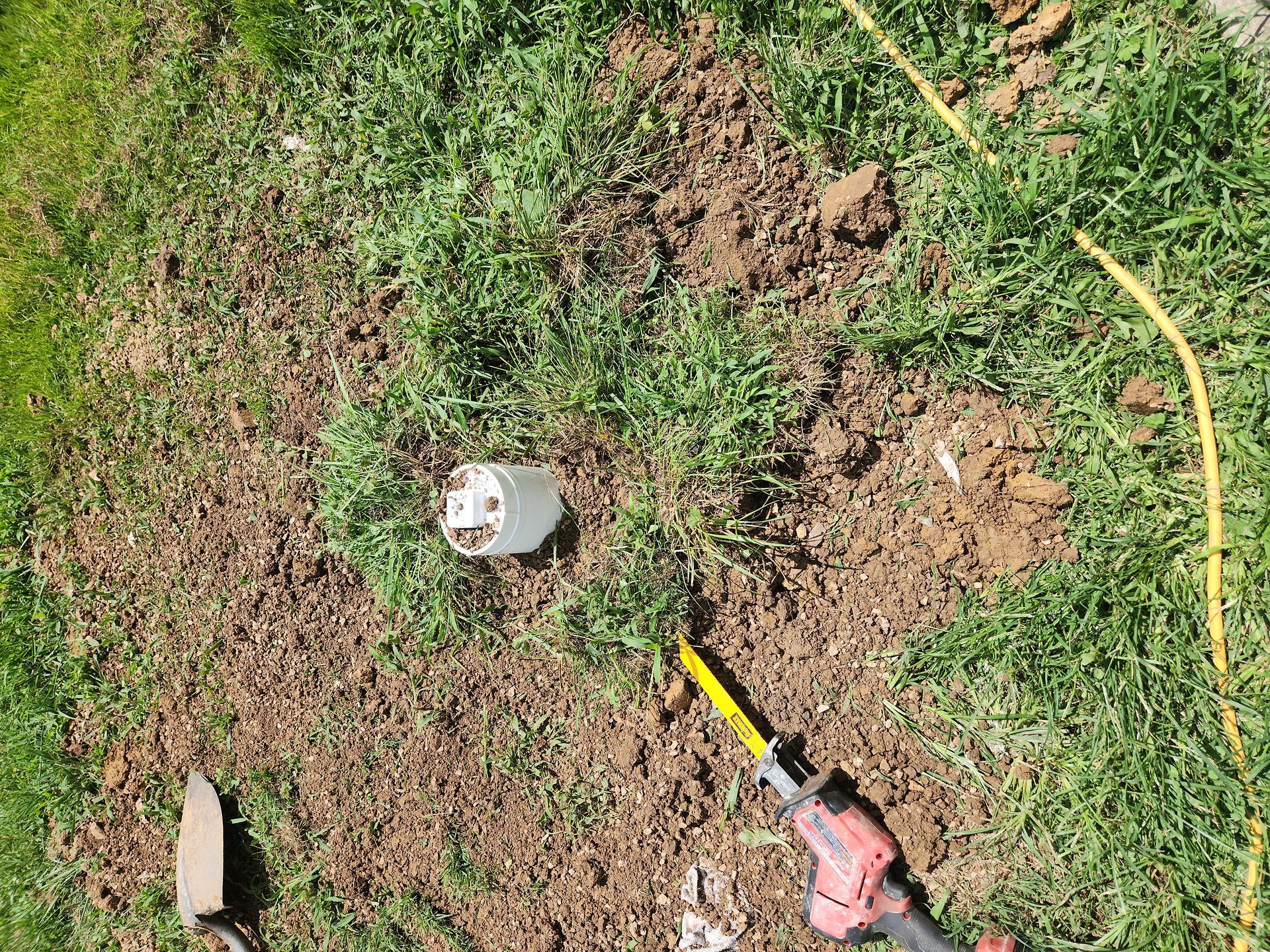 A yard hole with a white pipe, a red-and-yellow tool, and a yellow hose on patchy grass.