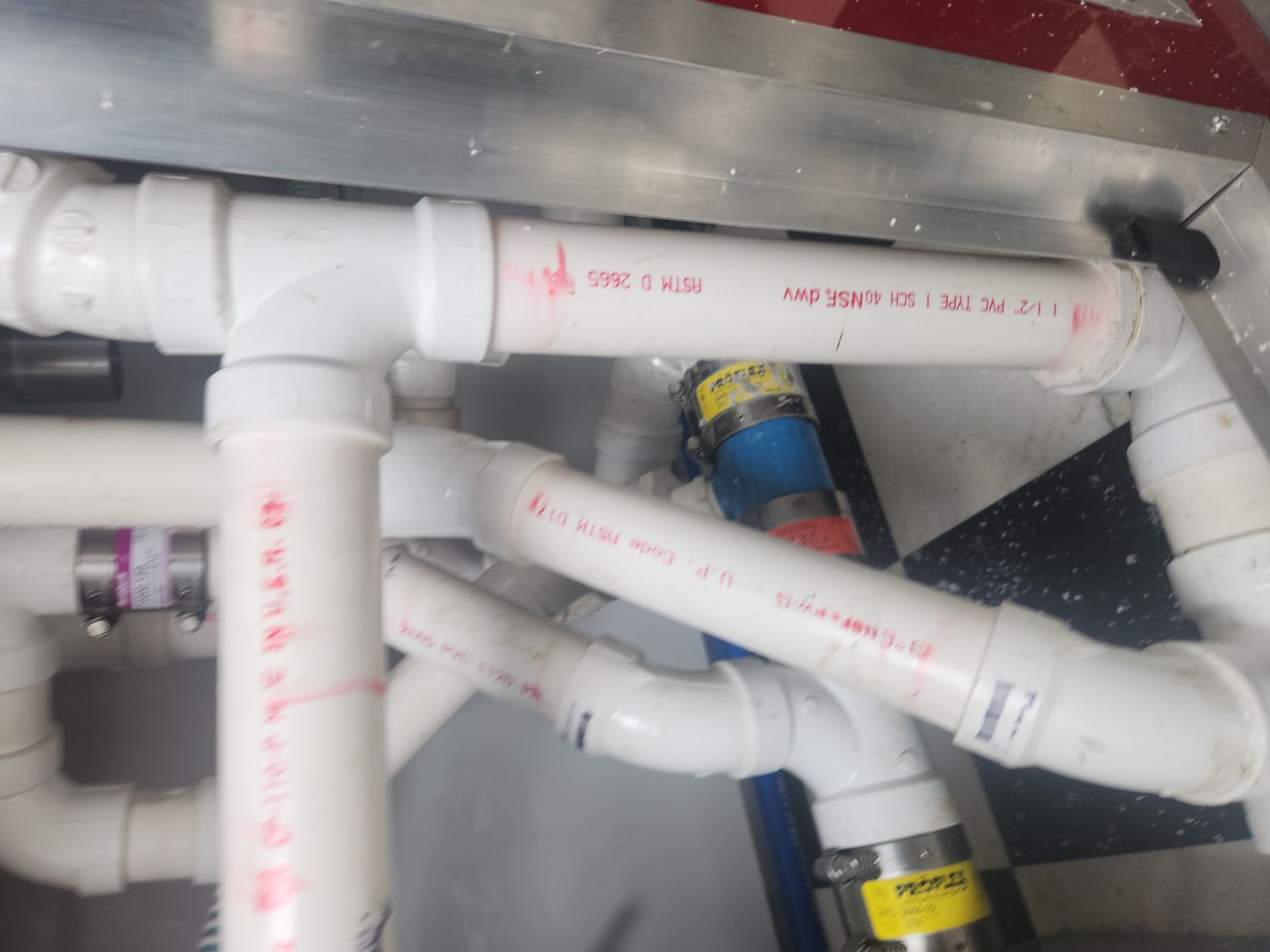 White PVC pipes with red markings in a cramped utility area, with a blue valve and yellow label.