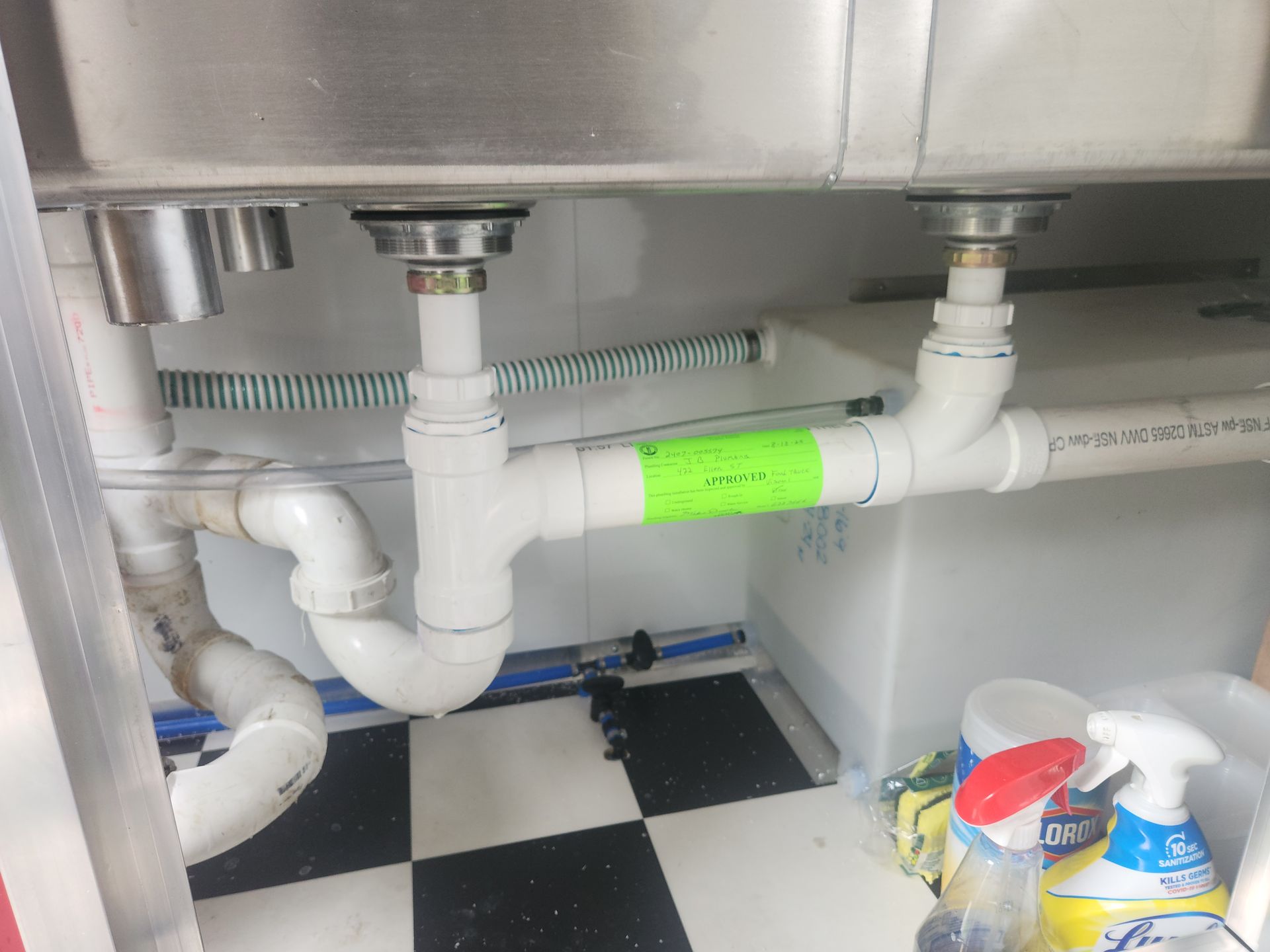 Under-sink plumbing with white PVC pipes and green tape beneath a stainless steel counter, on a black-and-white tile floor