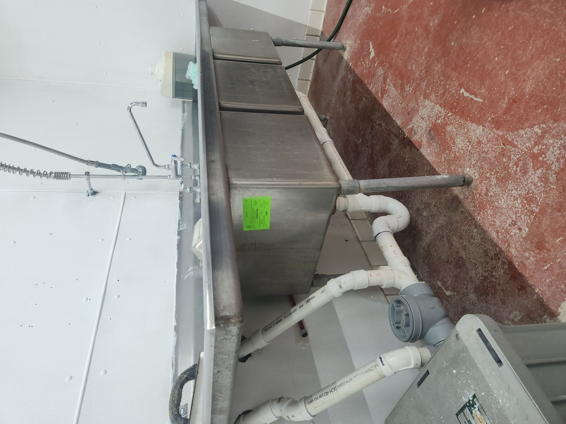 Stainless steel industrial sink with white pipes and a green label against a white and red wall.