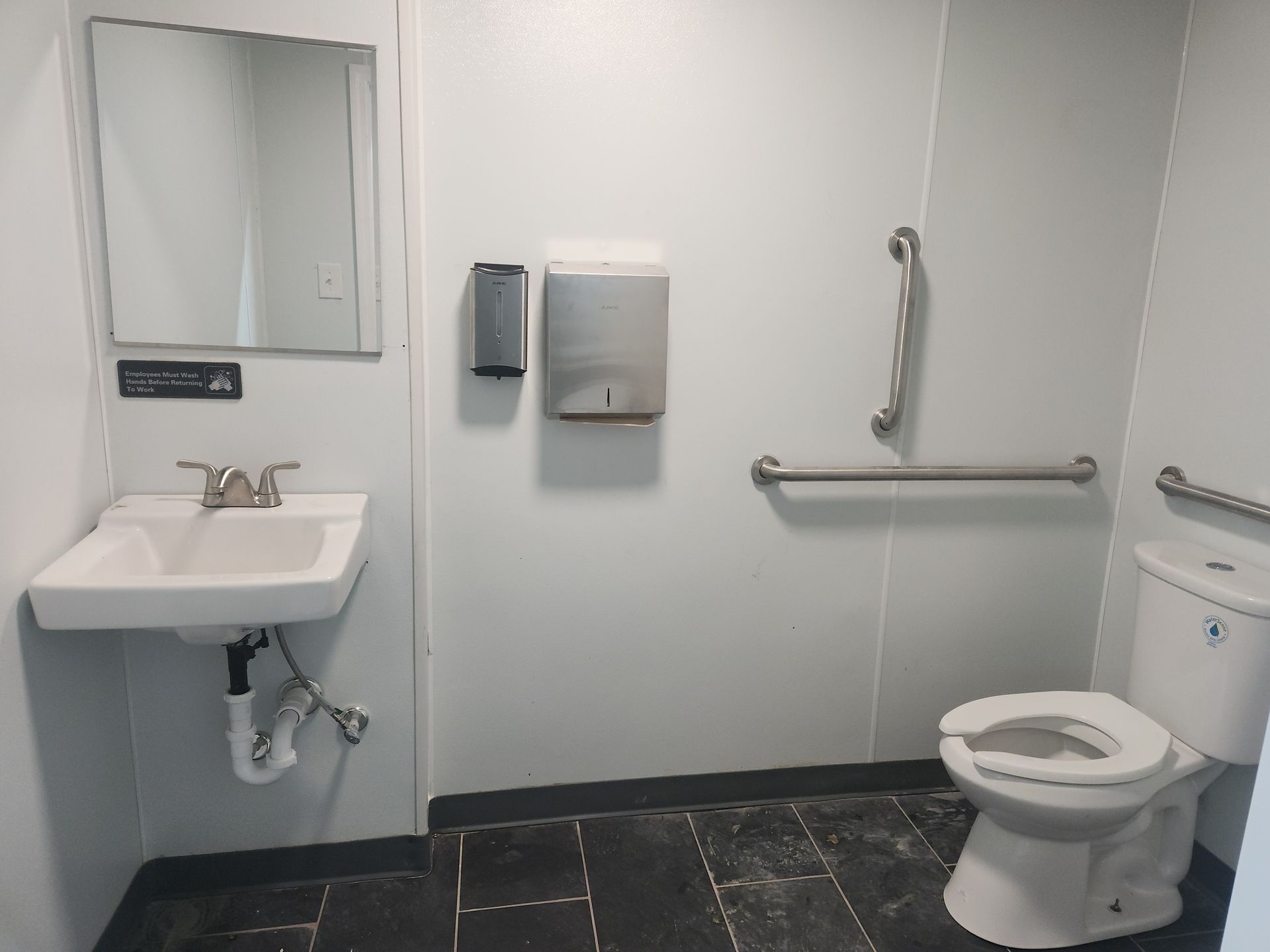 Accessible restroom with sink, mirror, grab bars, and toilet on dark floor tiles