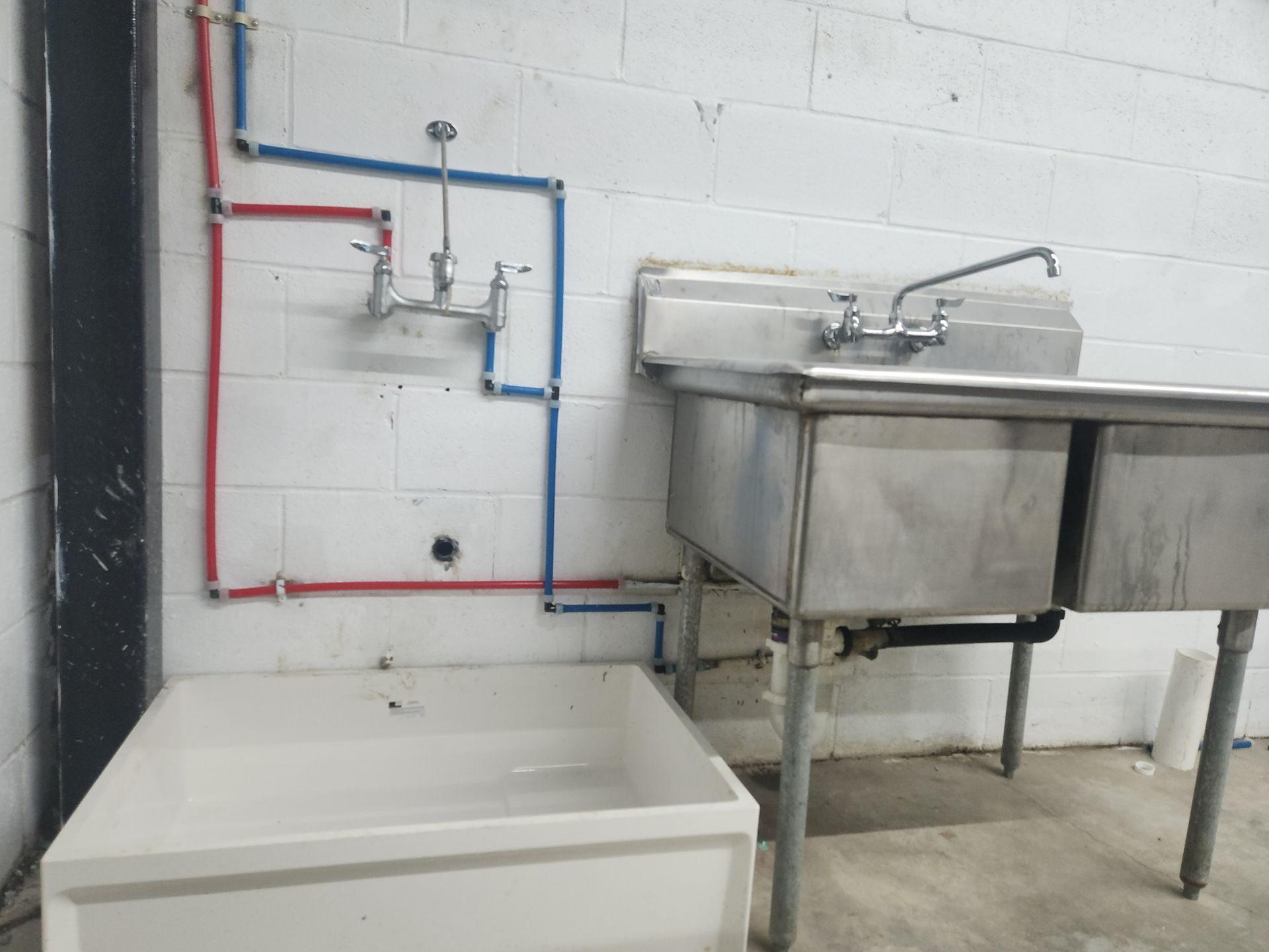 Utility sink and stainless-steel basin with exposed red and blue plumbing against a white cinder block wall