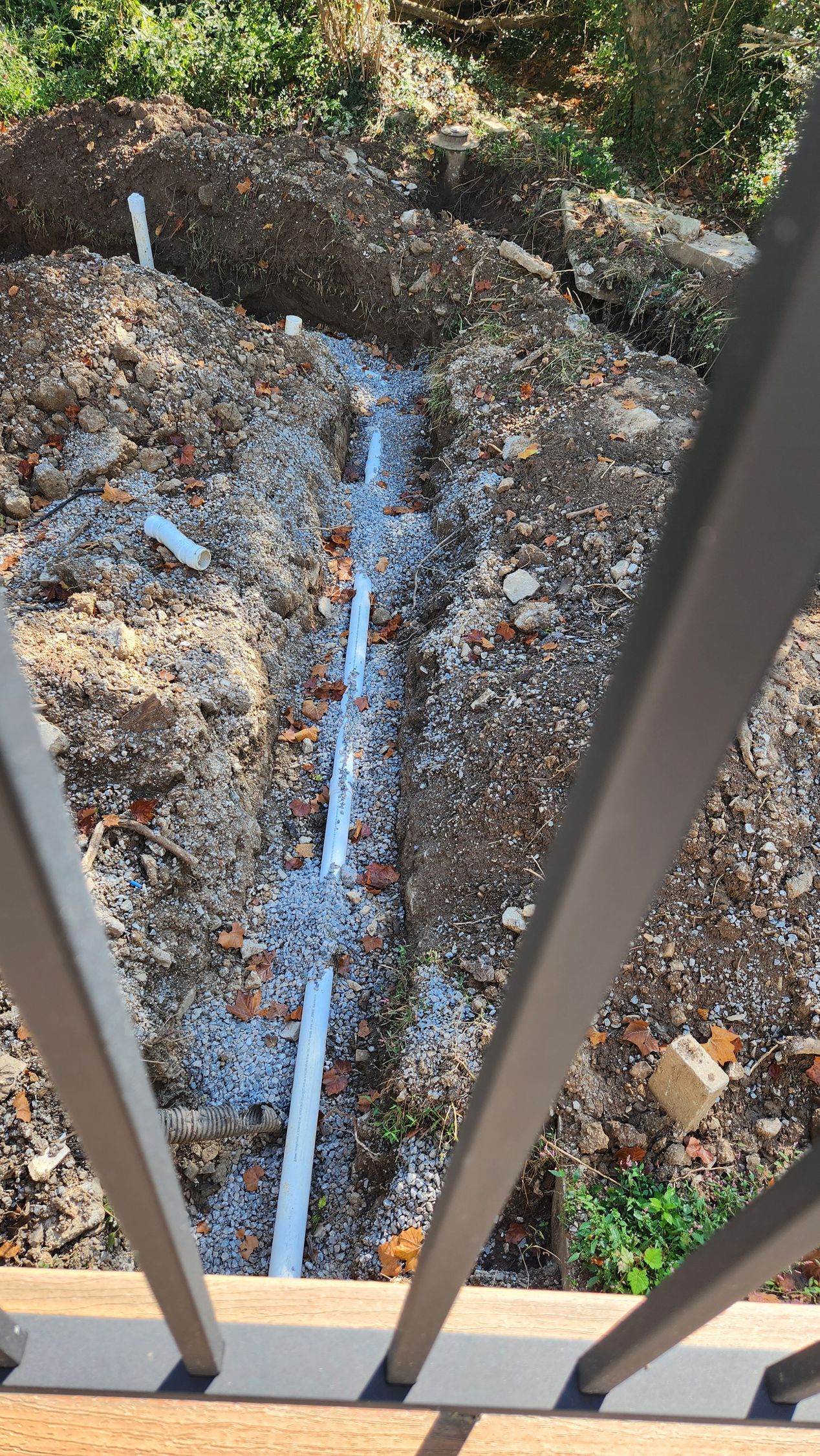 View from a balcony railing onto a rocky, debris-filled ground with a narrow drainage pipe or channel below