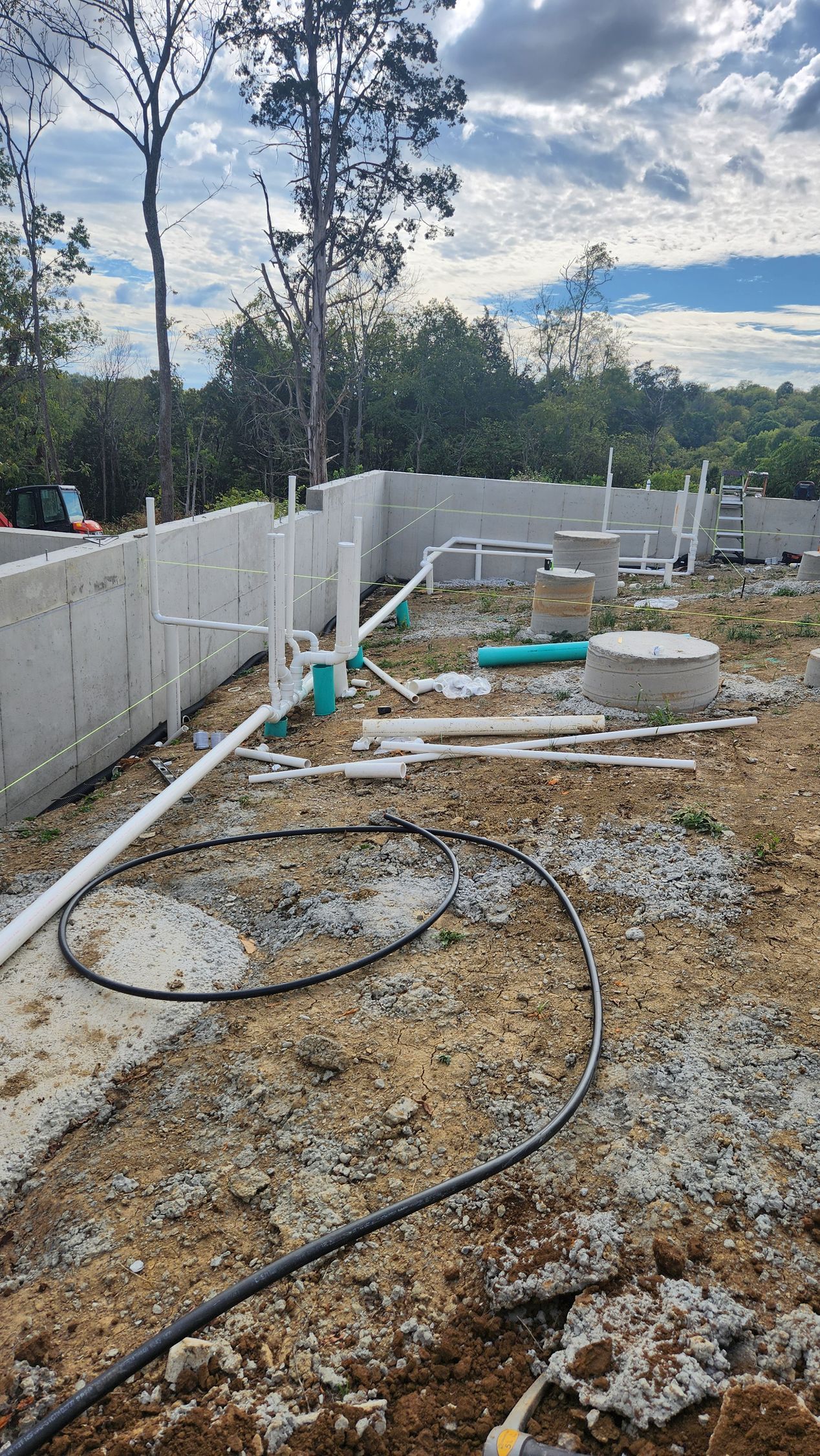 Construction site with white fence, muddy ground, scattered pipes, and tall trees in the background