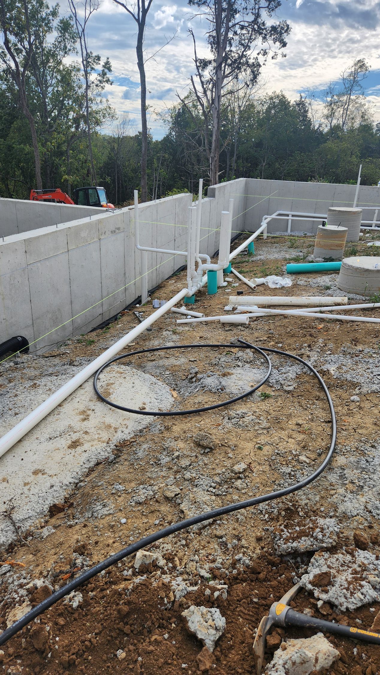Concrete foundation under construction with pipes, gravel, and a gray retaining wall in a wooded area.