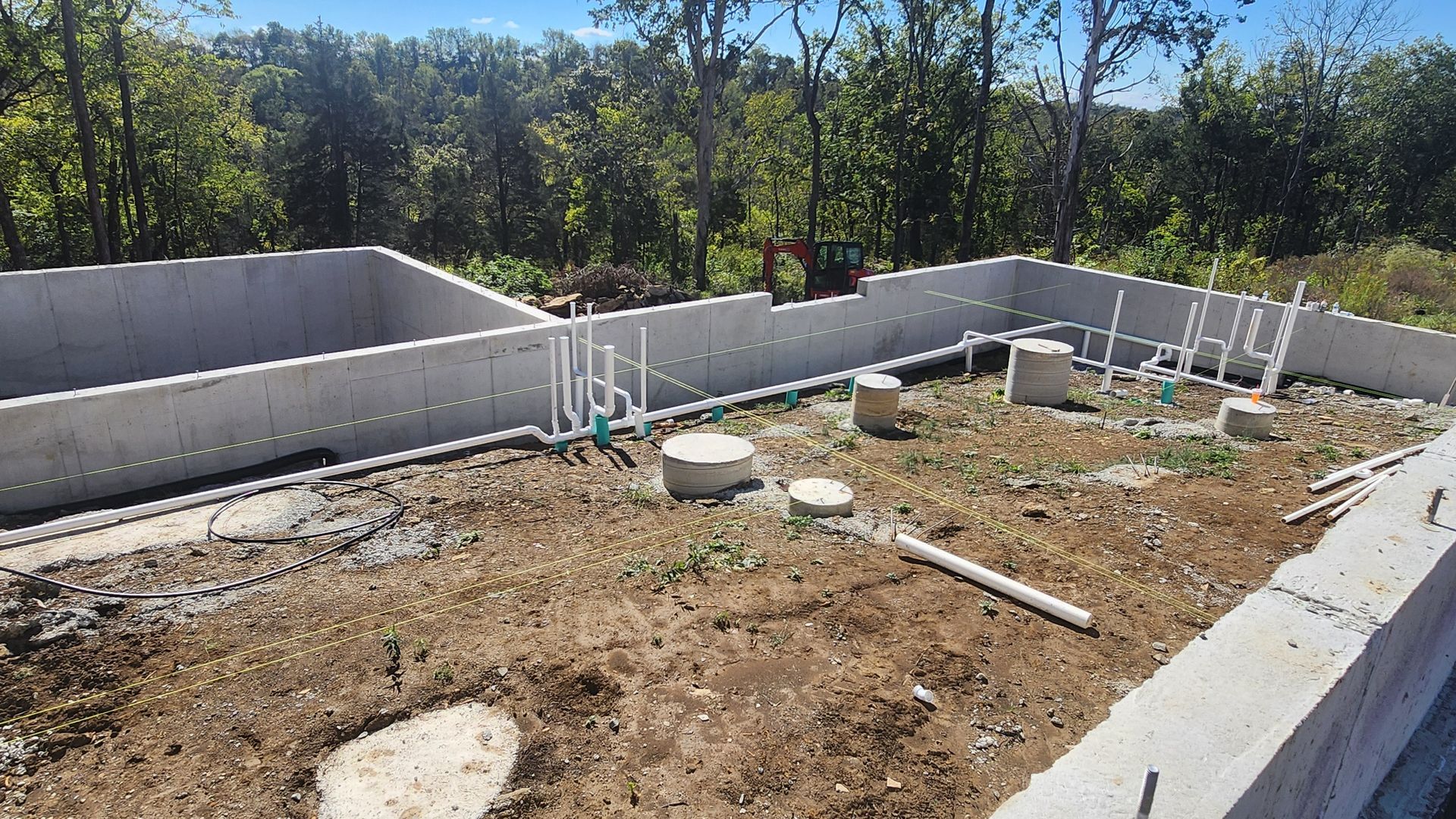 Partially built concrete foundation in a wooded outdoor lot with pipes and construction materials scattered around