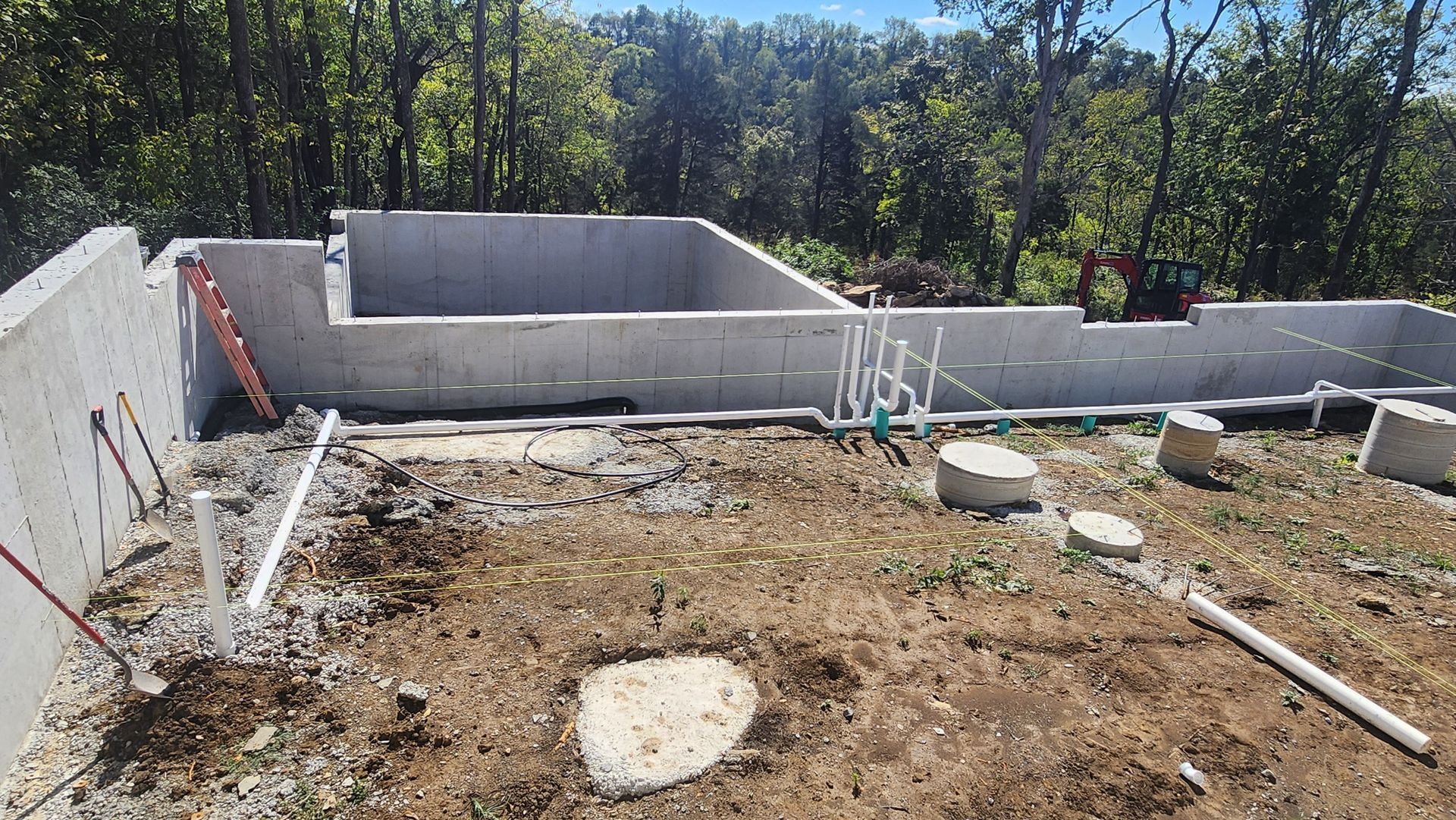 Partially built concrete foundation on a dirt lot with construction materials and trees in the background