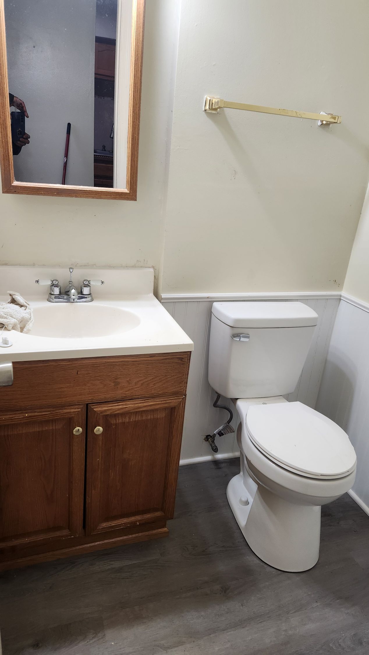 Small bathroom with white toilet, wood vanity, mirror, towel bar, and gray flooring.