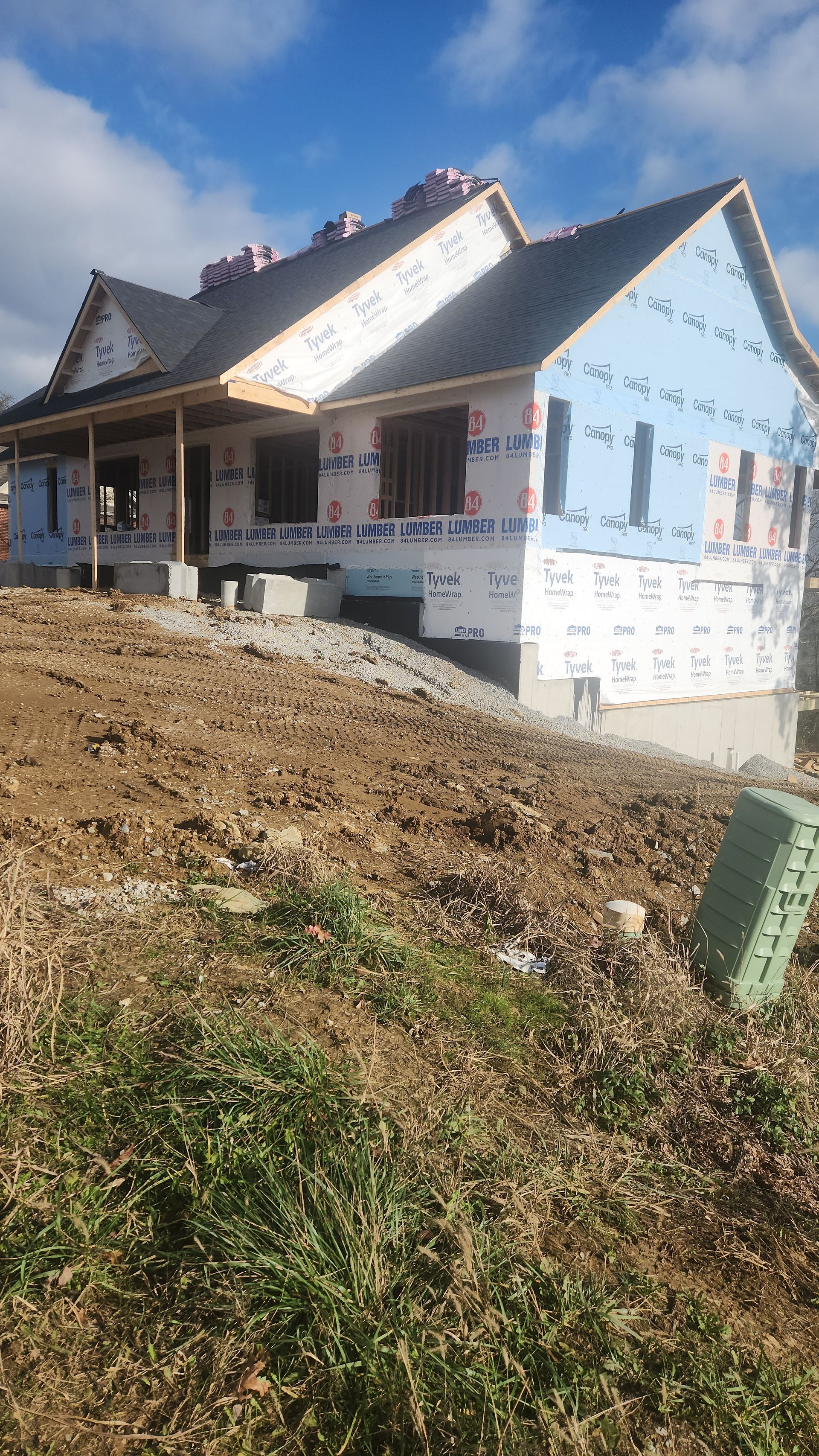 Partially built house with blue siding wrap and steep roof on a dirt lot under a cloudy sky