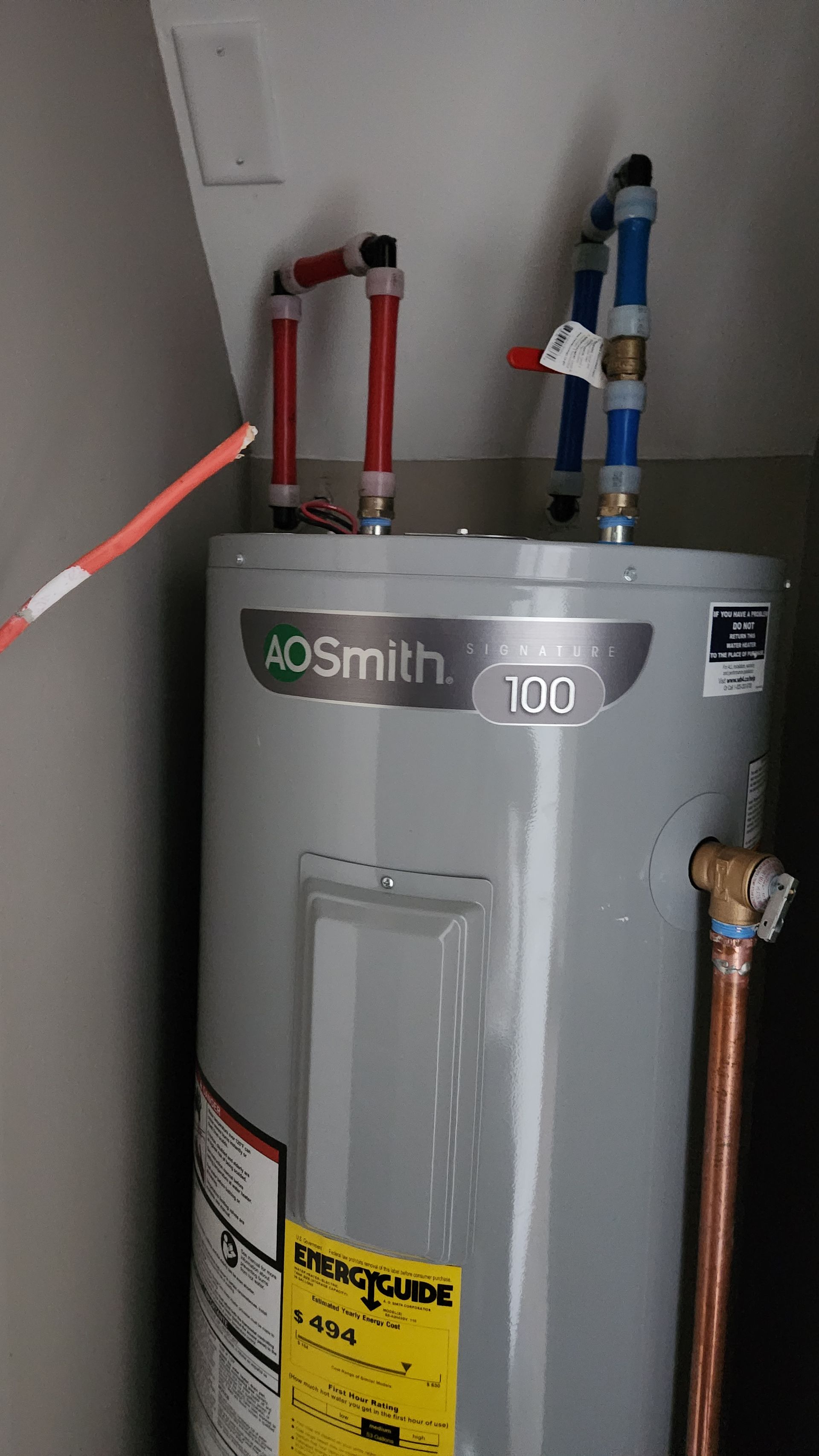 Water heater with red and blue pipes in a utility closet; a hand points toward it.