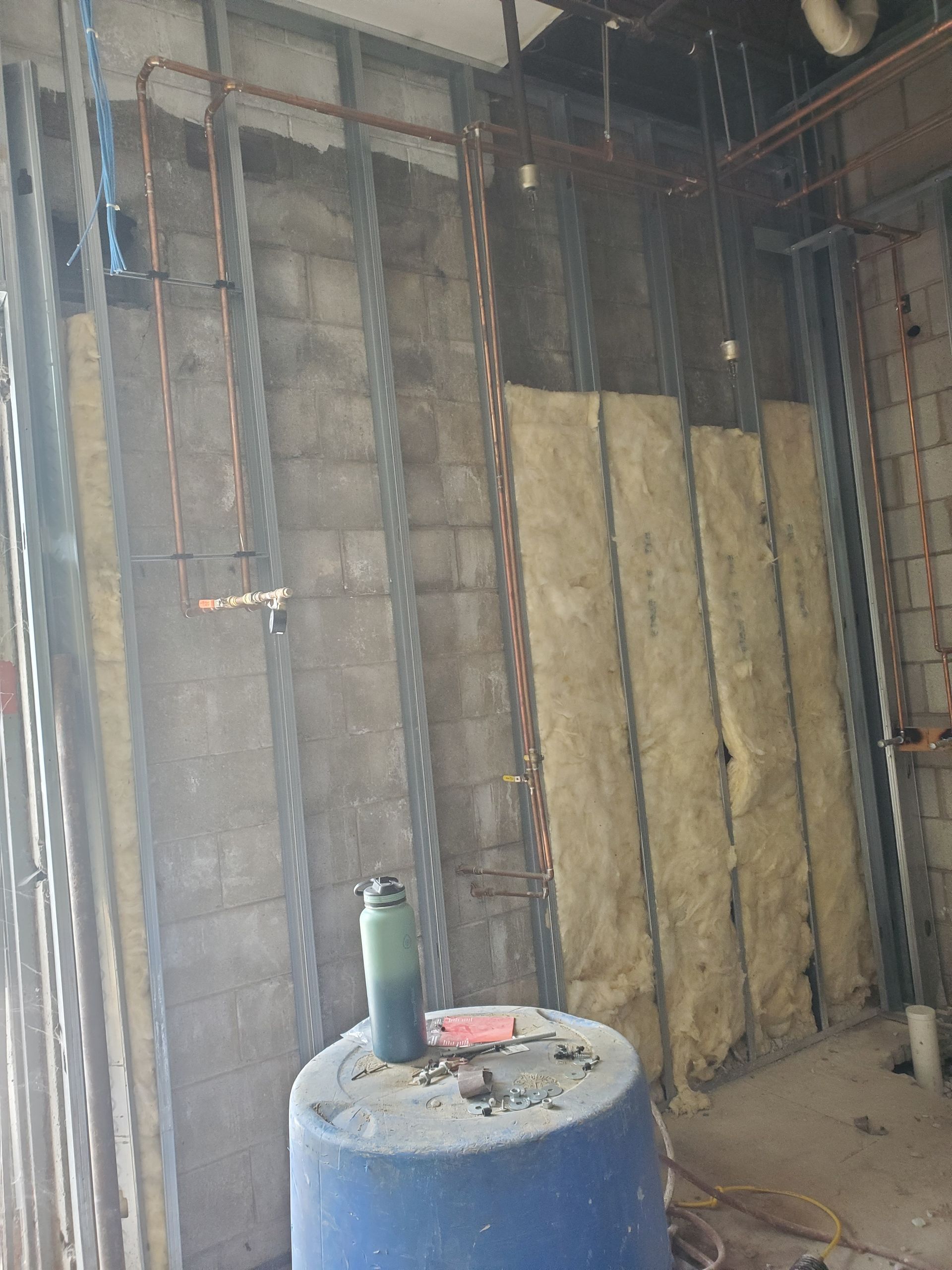Unfinished interior wall framing with exposed insulation and a blue tank in the foreground.