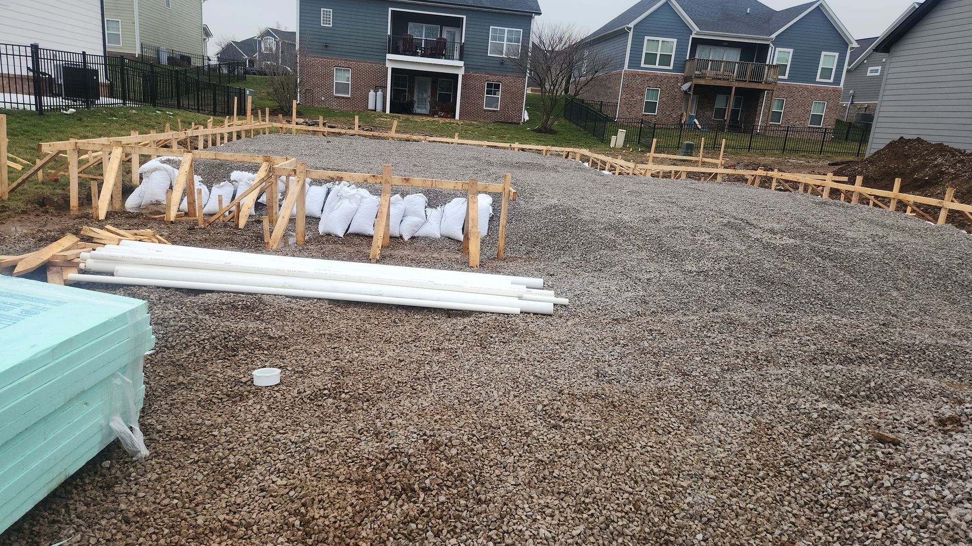 Construction site with gravel foundation, lumber stacks, and houses in the background
