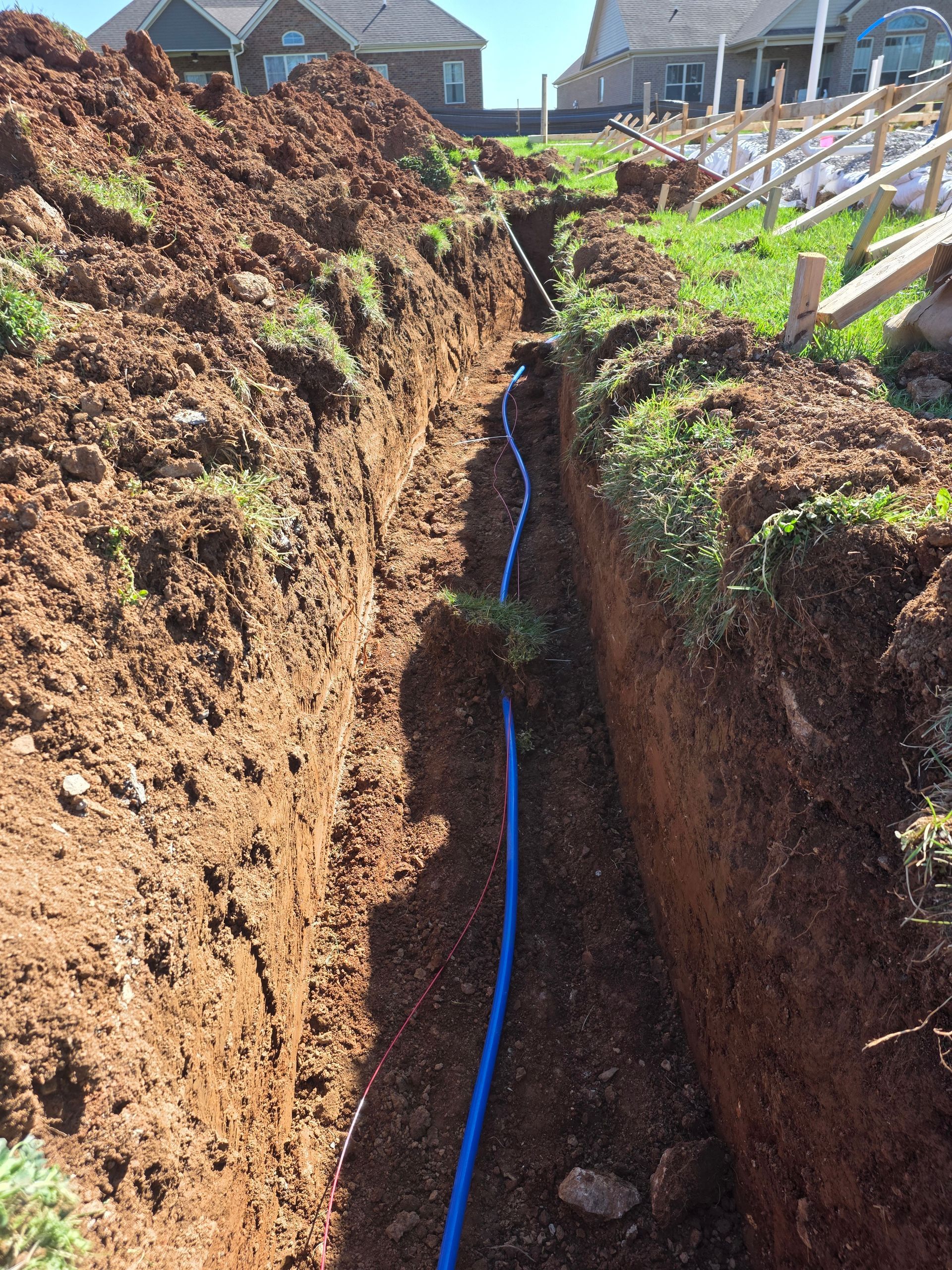 Deep trench in a backyard with a blue utility cable running along the bottom beside houses and fences