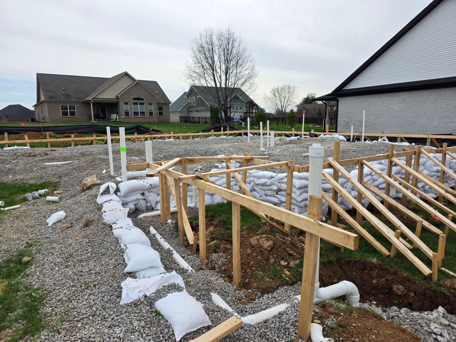 Foundation site with wooden framing and white pipes in a gravel lot near houses under an overcast sky