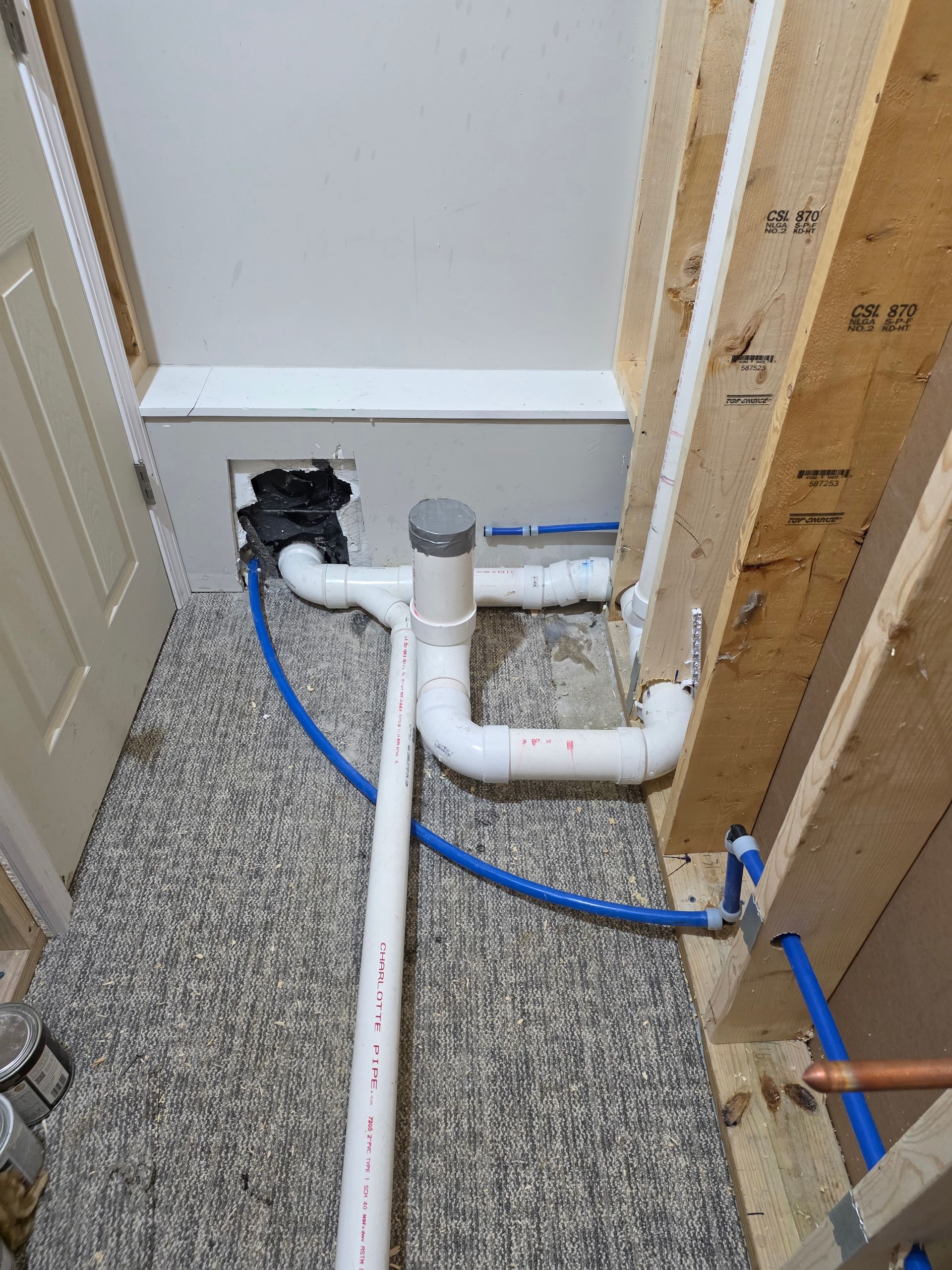 Exposed white PVC plumbing pipes and blue cable in a narrow unfinished utility nook, beside a door and wall studs