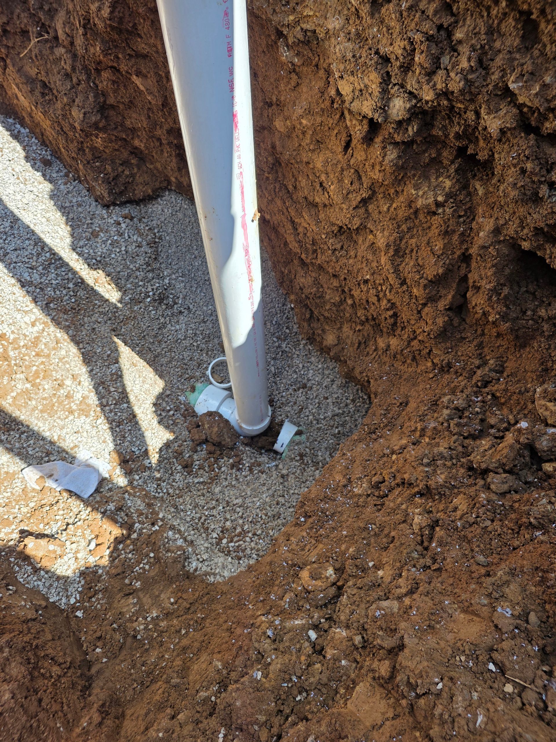 White pipe emerging from gravel beside a dirt trench or hole outdoors.
