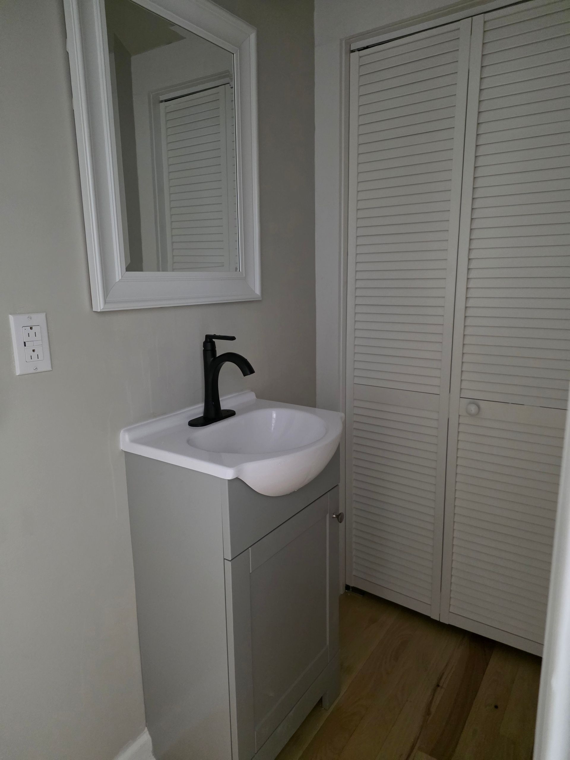 Small bathroom vanity with black faucet, mirror, and white louvered closet doors.