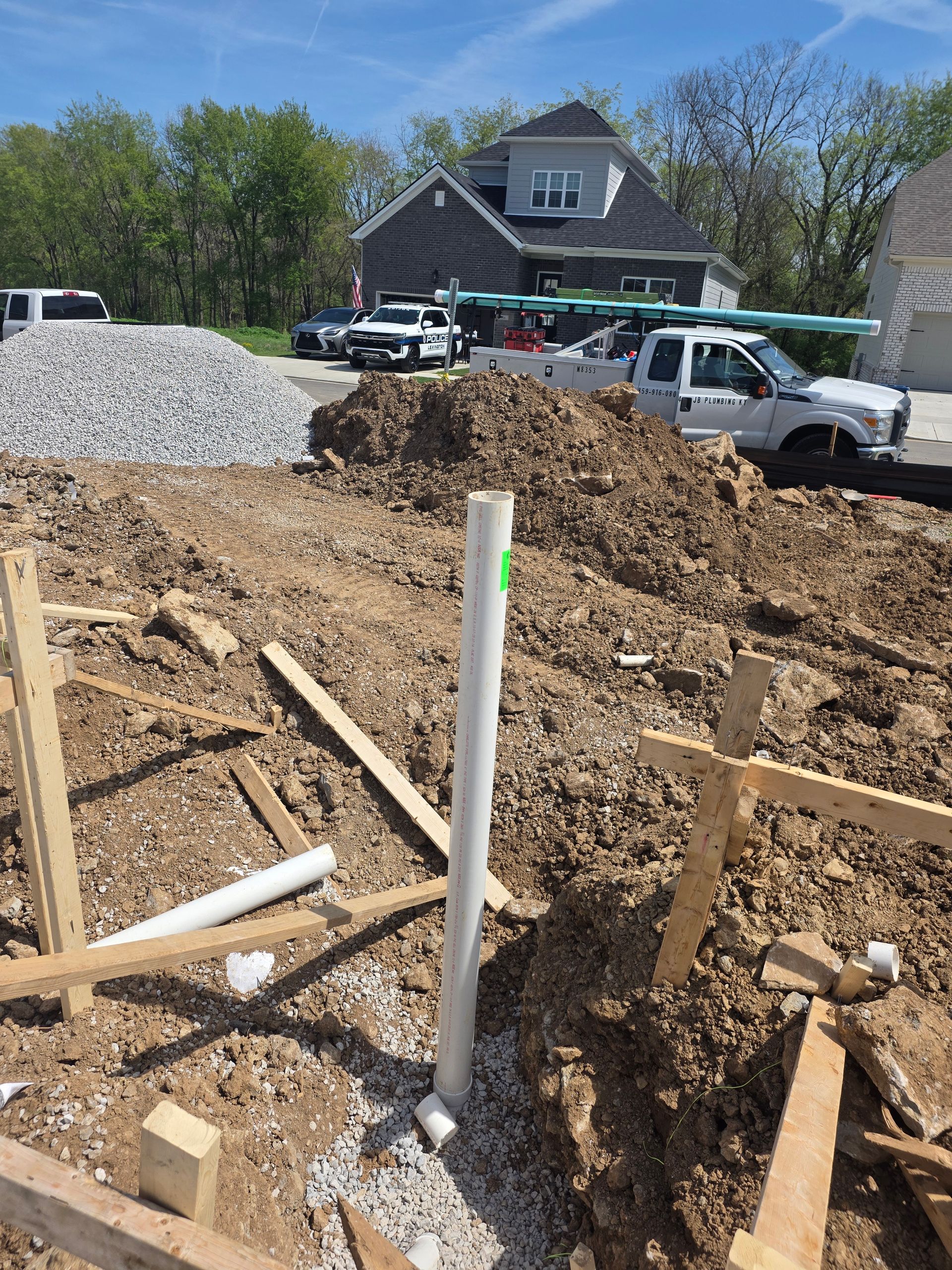 Construction trench with white pipe and wooden stakes beside a gravel pile and parked cars.