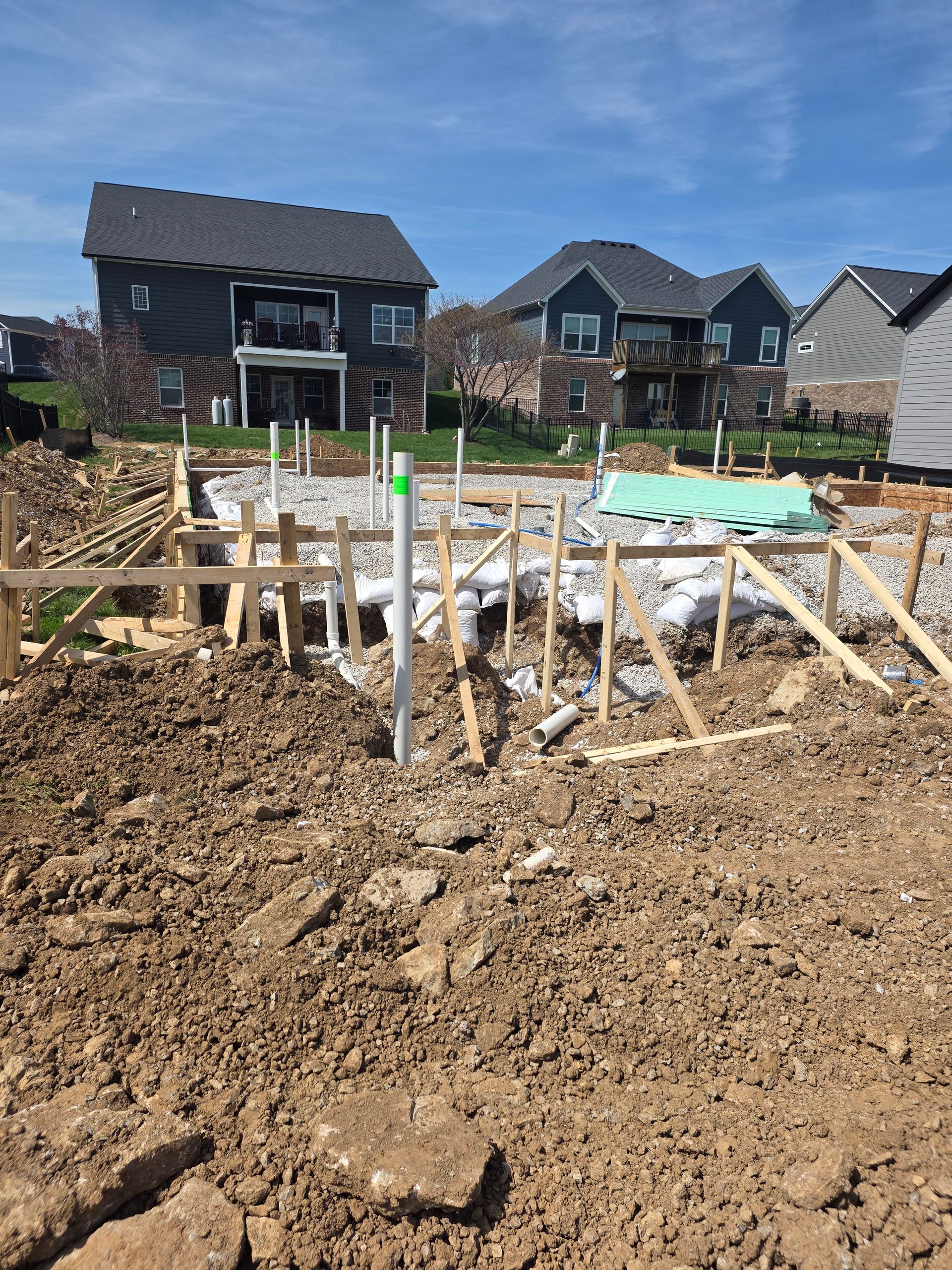 Residential construction site with rebar foundation forms in a muddy lot, houses in the background