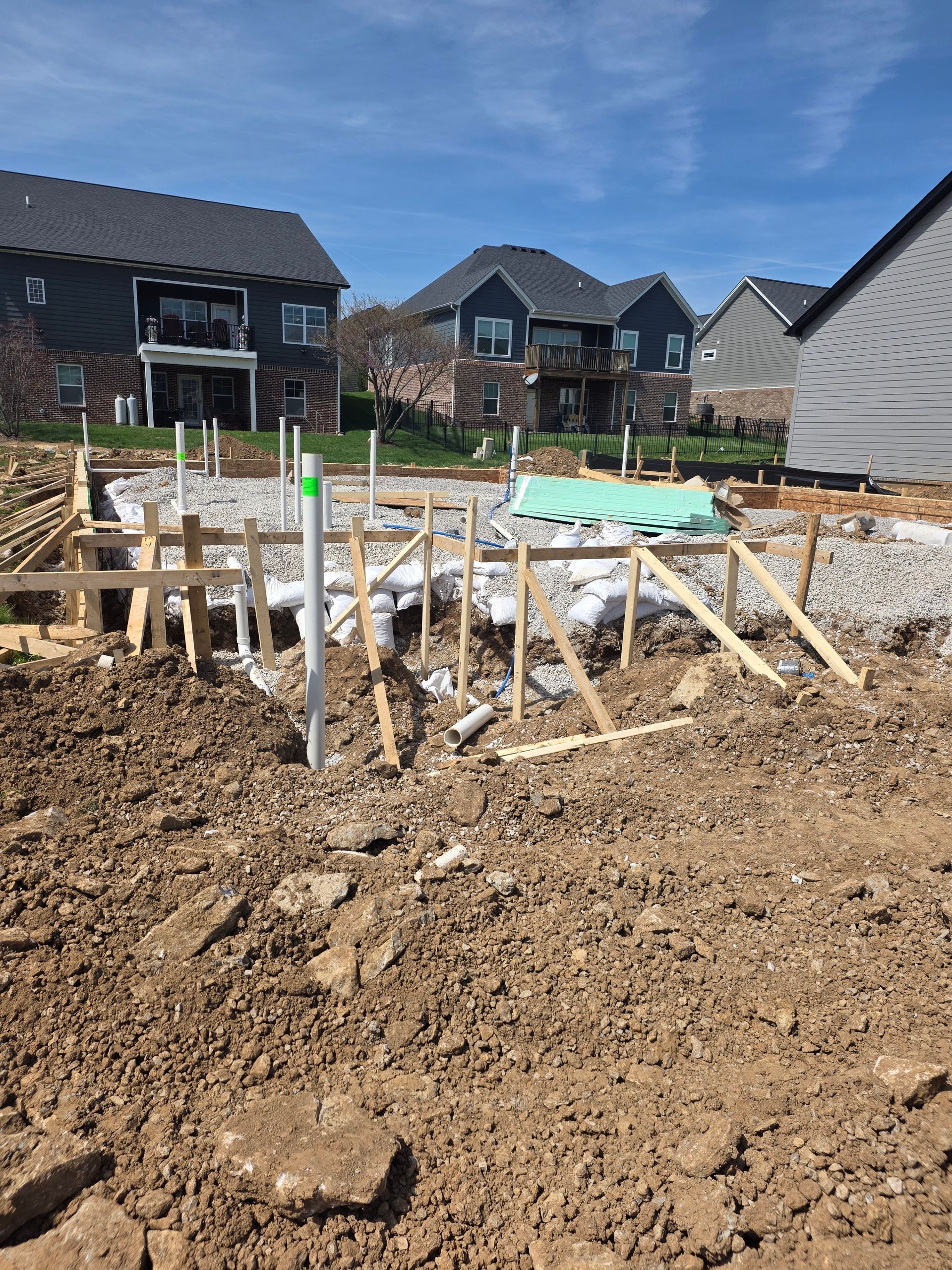 Construction site with foundation forms and rebar in a dirt lot, houses in the background