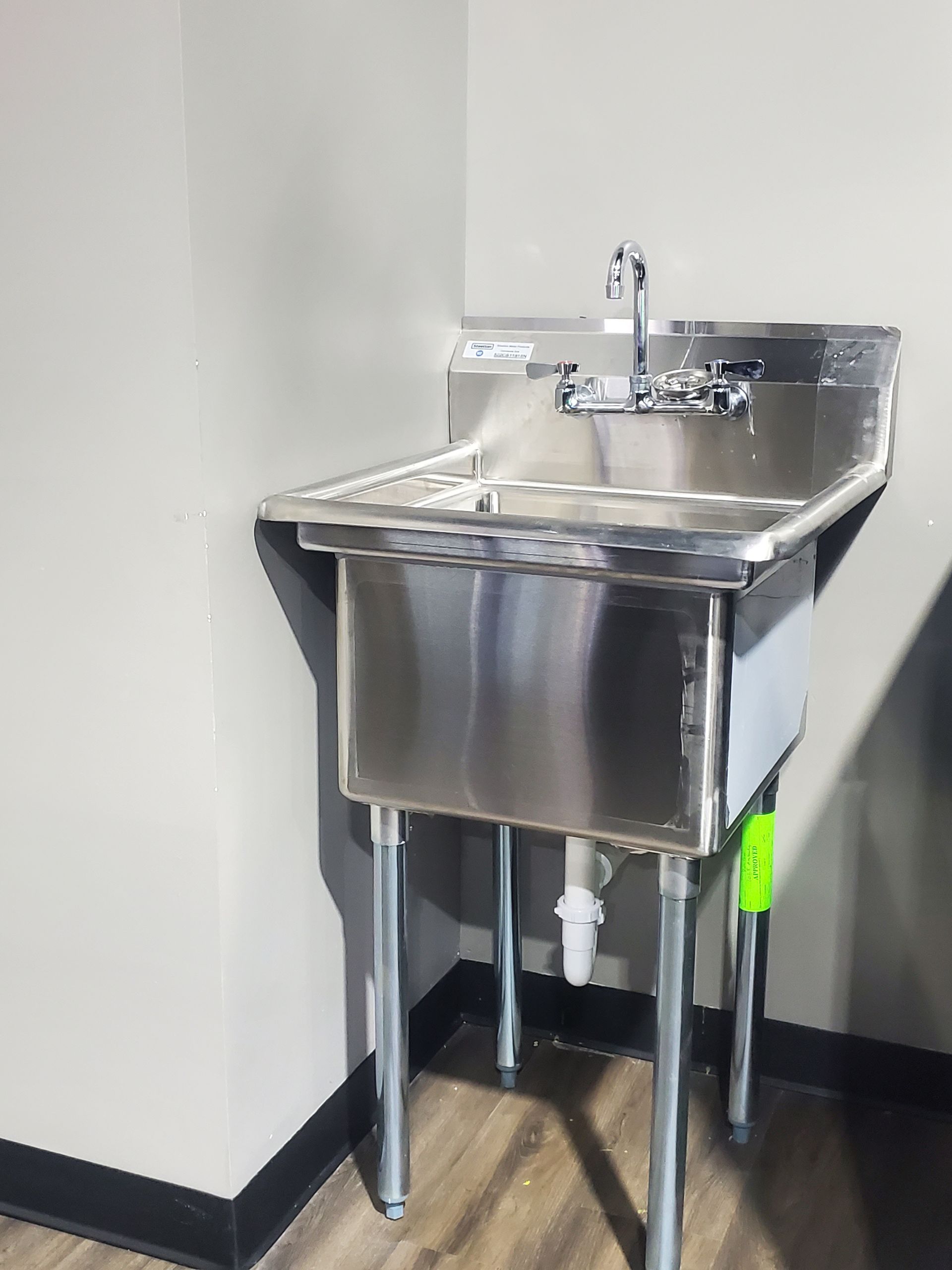 Stainless steel sink in a corner with faucet and spray hose, mounted in a plain white utility room.