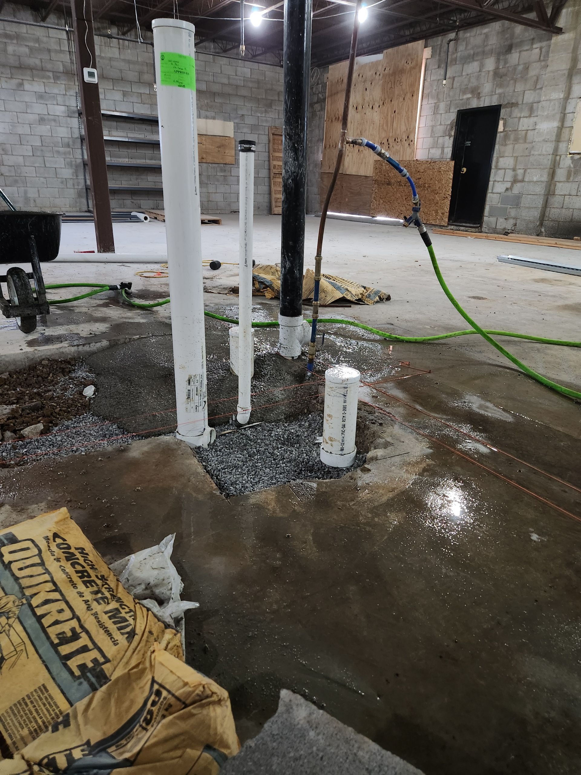 Indoor construction site with wet concrete floor, white pipes, debris, and exposed brick walls.