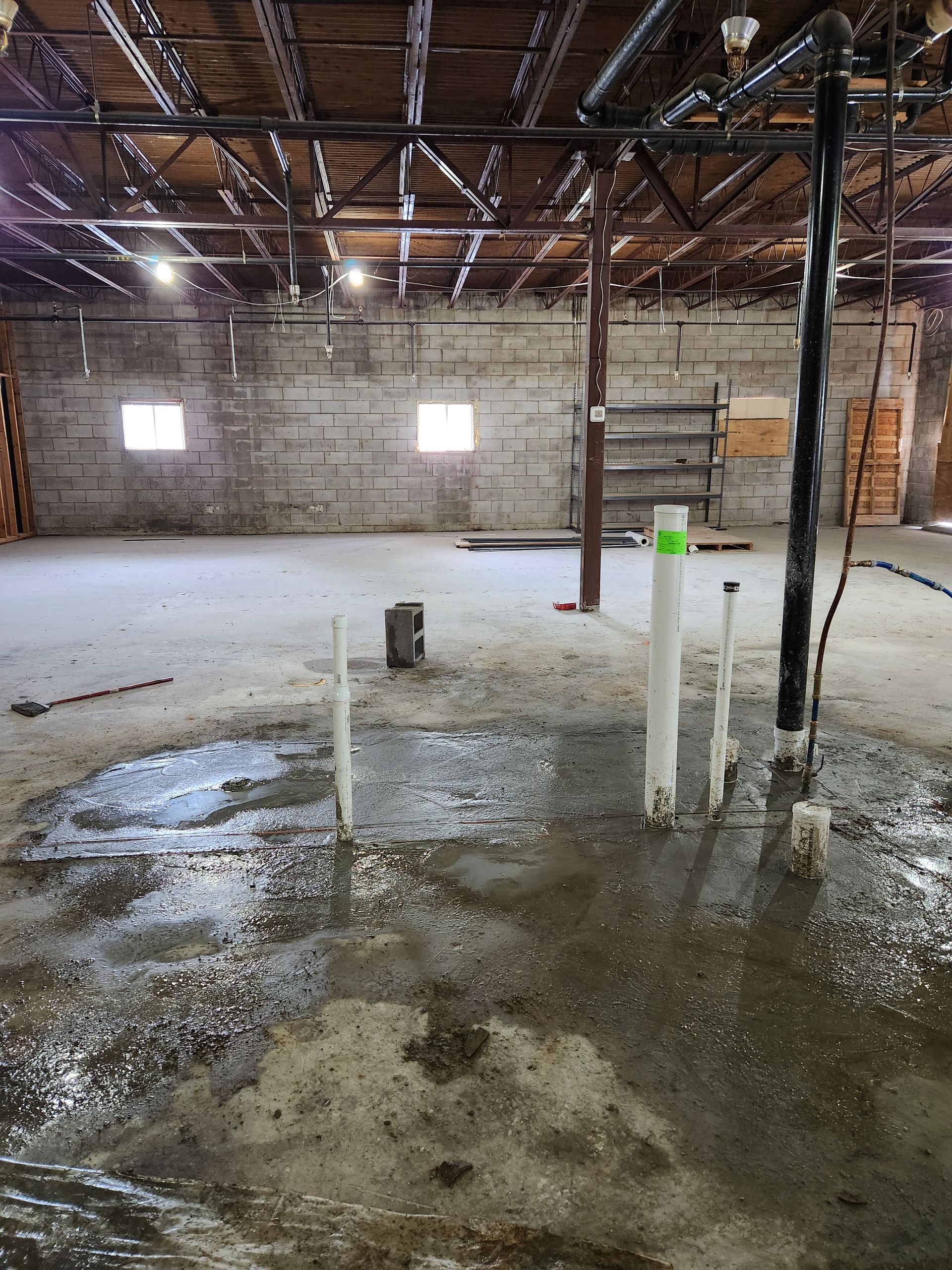 Abandoned warehouse interior with exposed beams, concrete floor, puddles, and white pipes near center