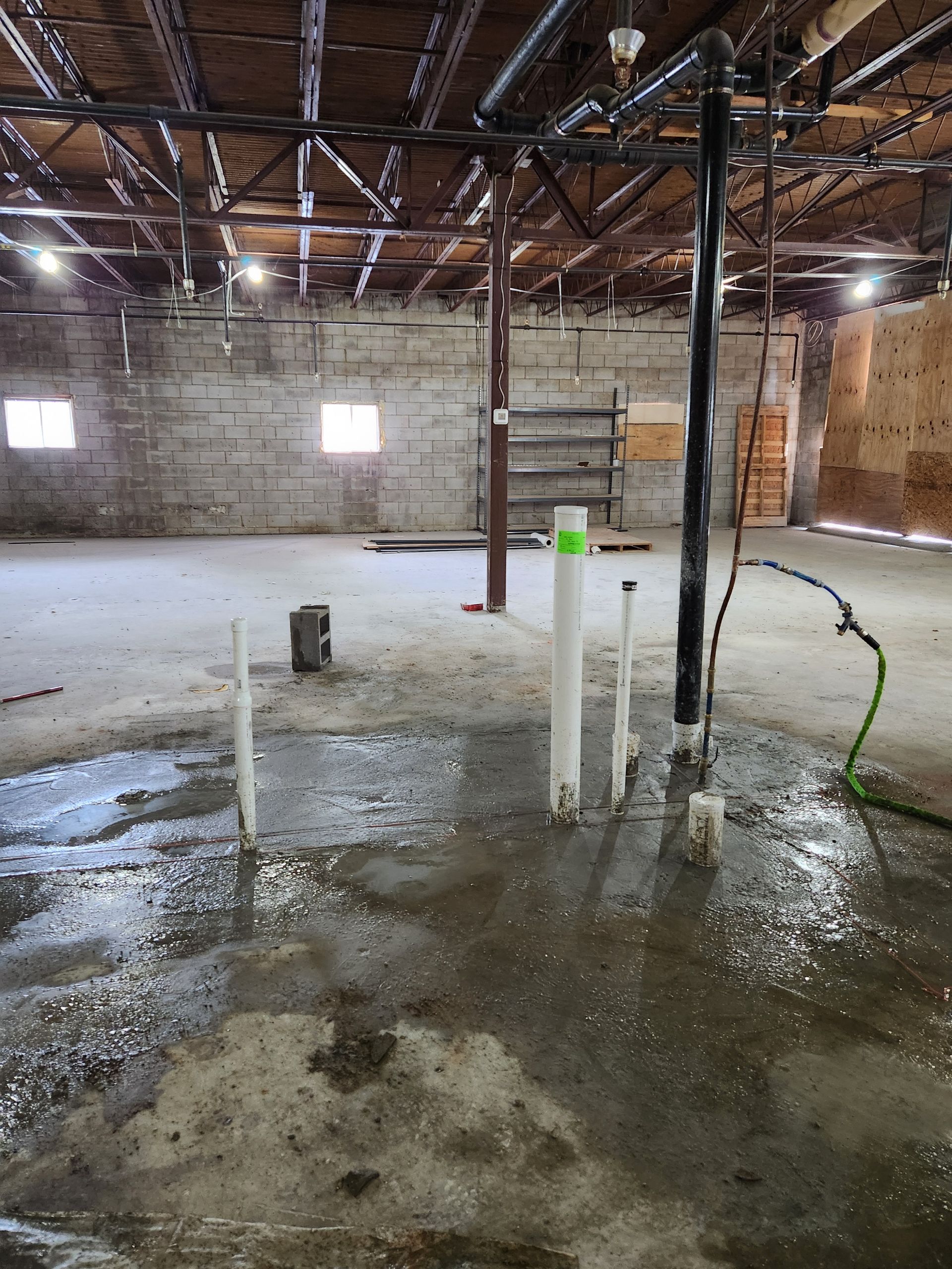 Basement under construction with wet concrete floor, exposed pipes, and unfinished brick walls.