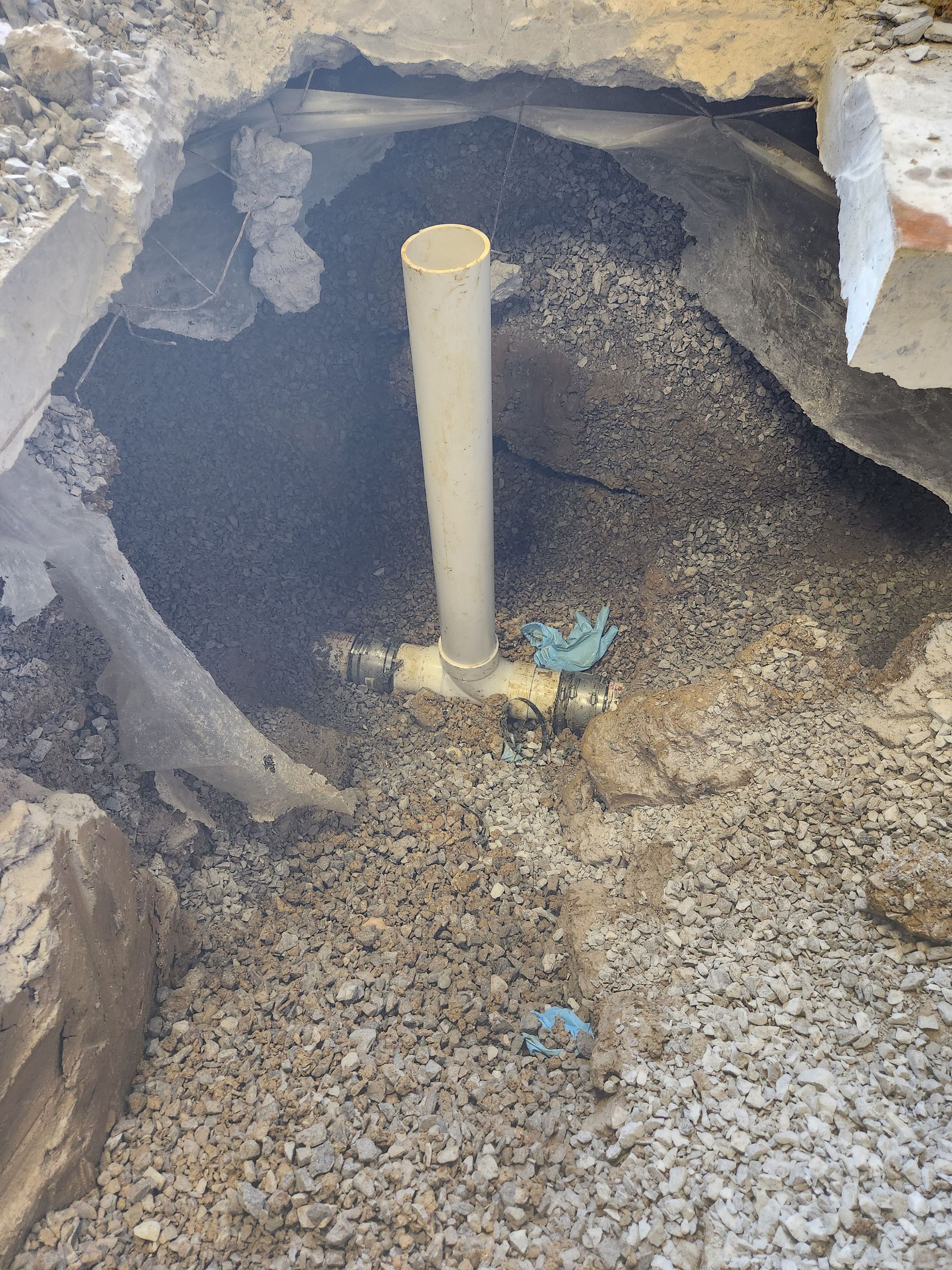Open excavation pit with a vertical PVC pipe and gravel around it, near a rough concrete wall.