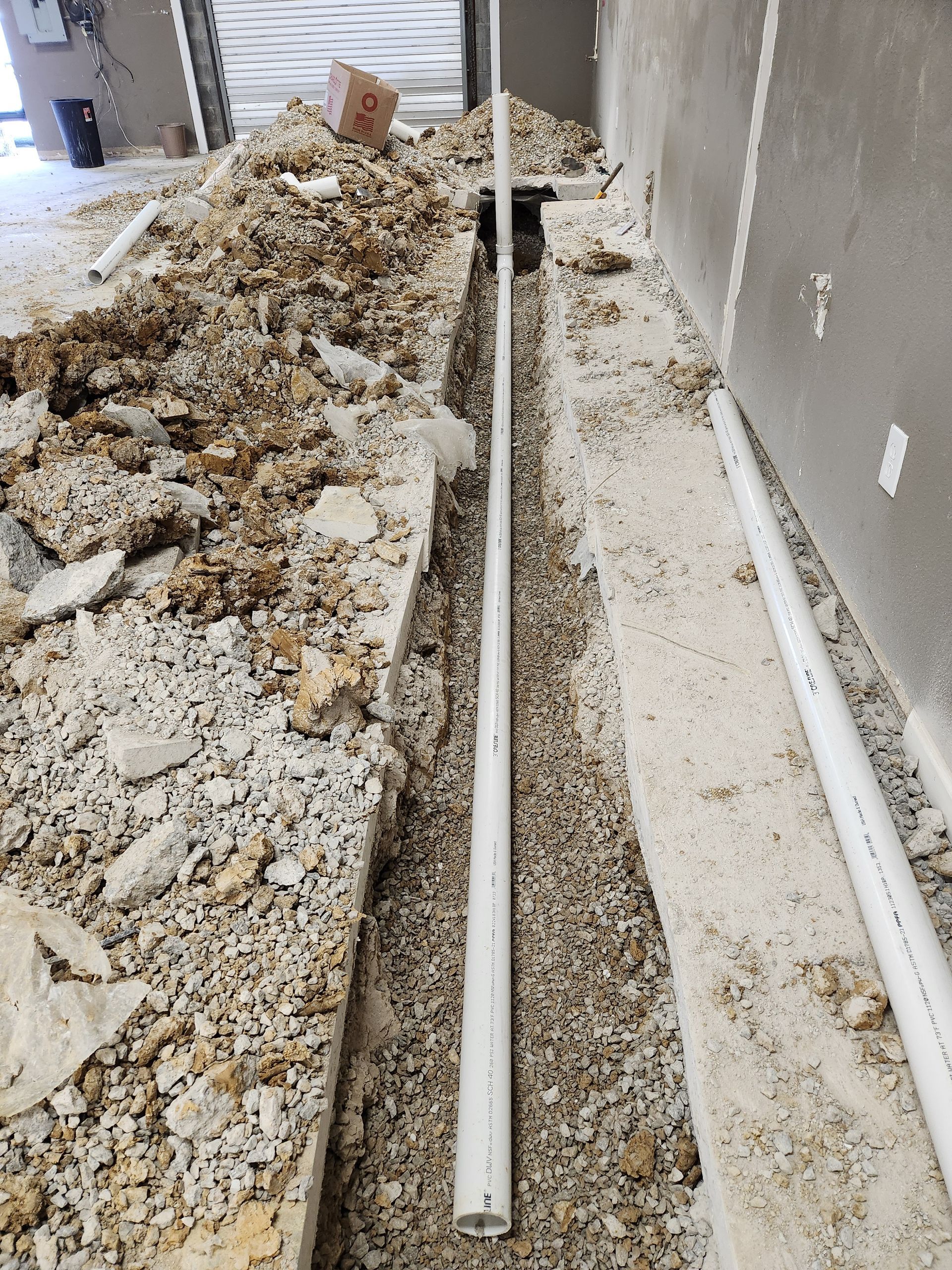 Debris-strewn construction trench with white PVC pipes beside a concrete wall