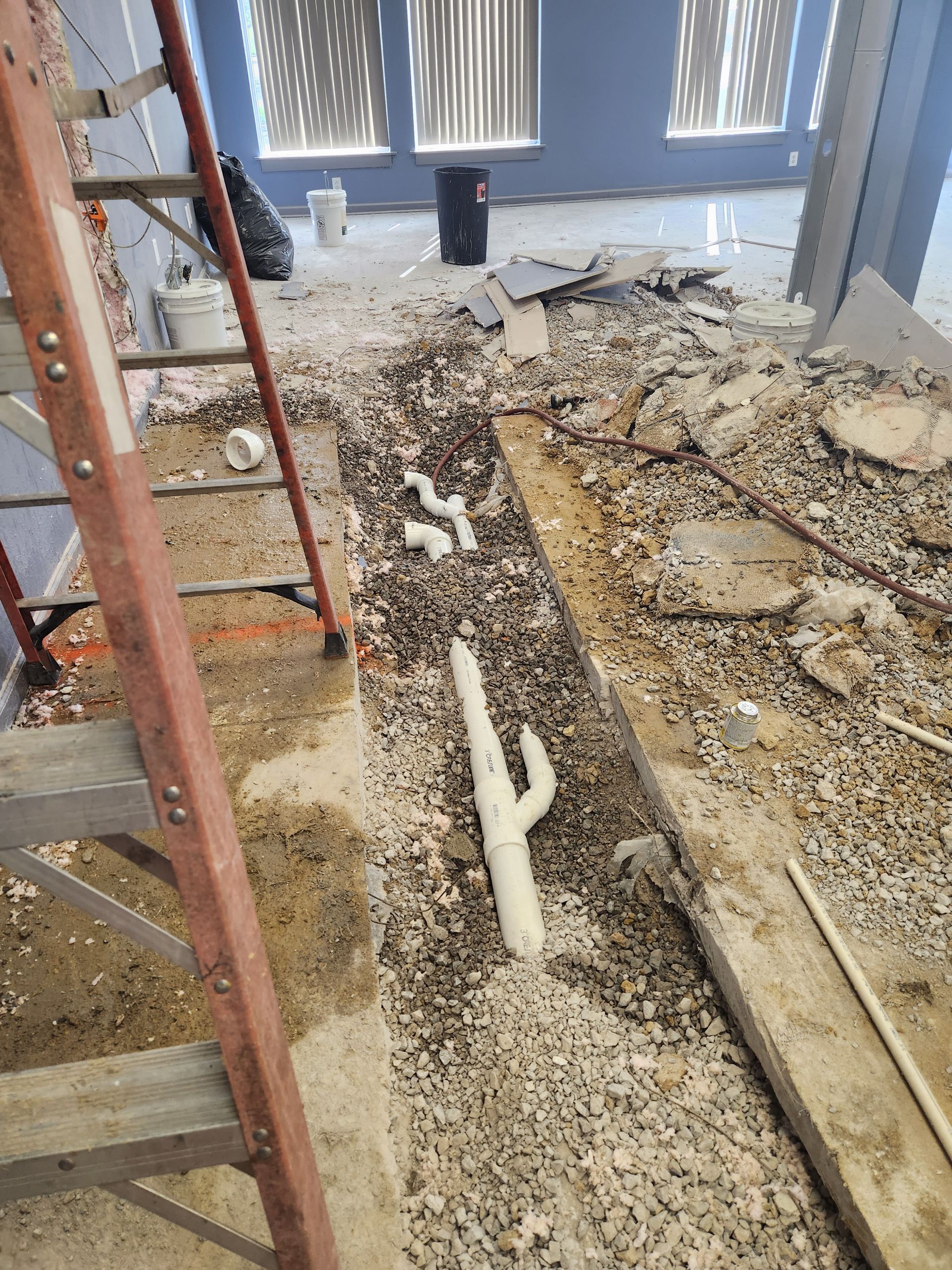 Construction debris scattered on a floor with exposed pipes, a ladder, and windowed walls.