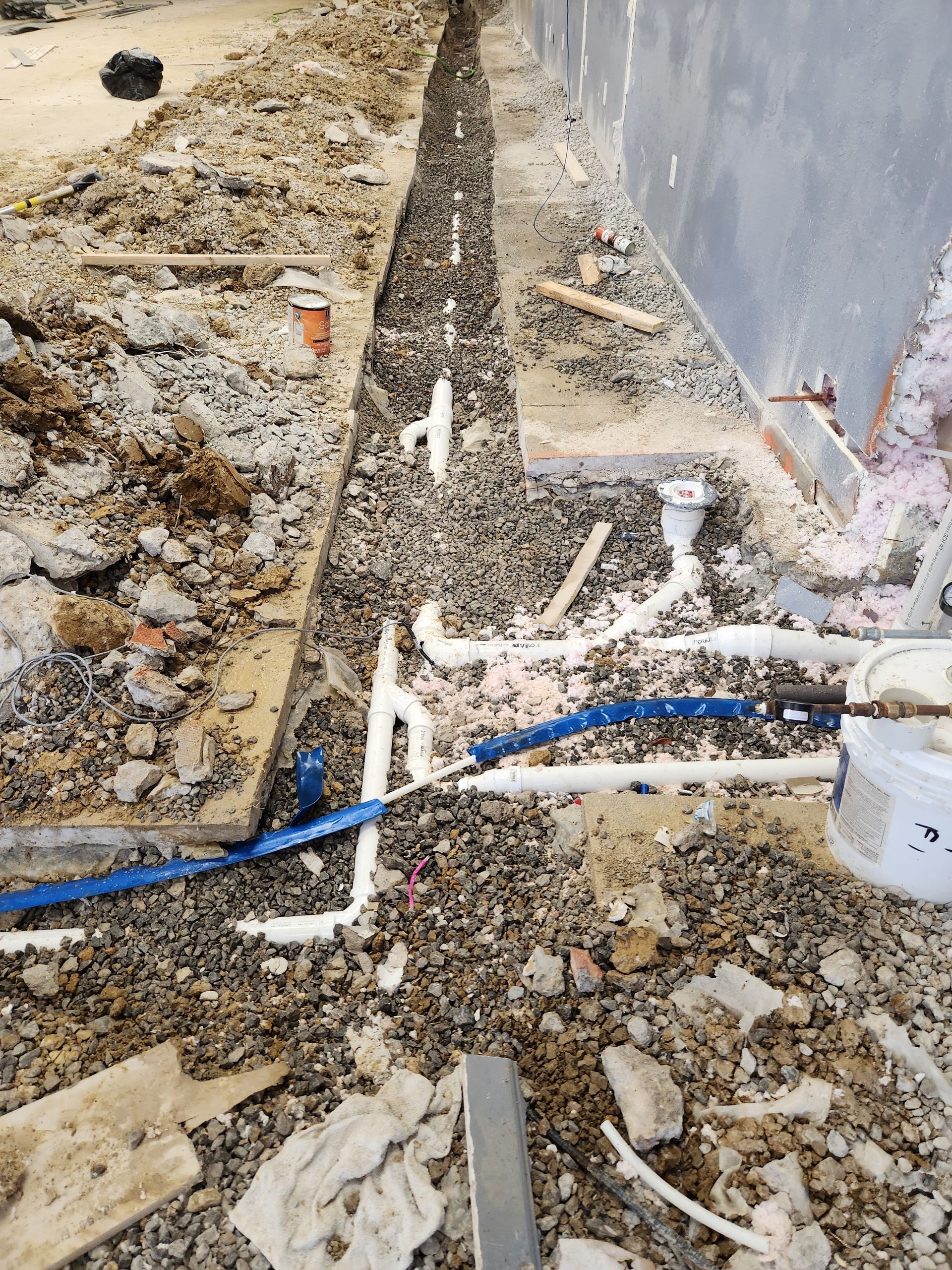 Construction debris beside a wall with exposed plumbing pipes and a blue hose along a rough trench.