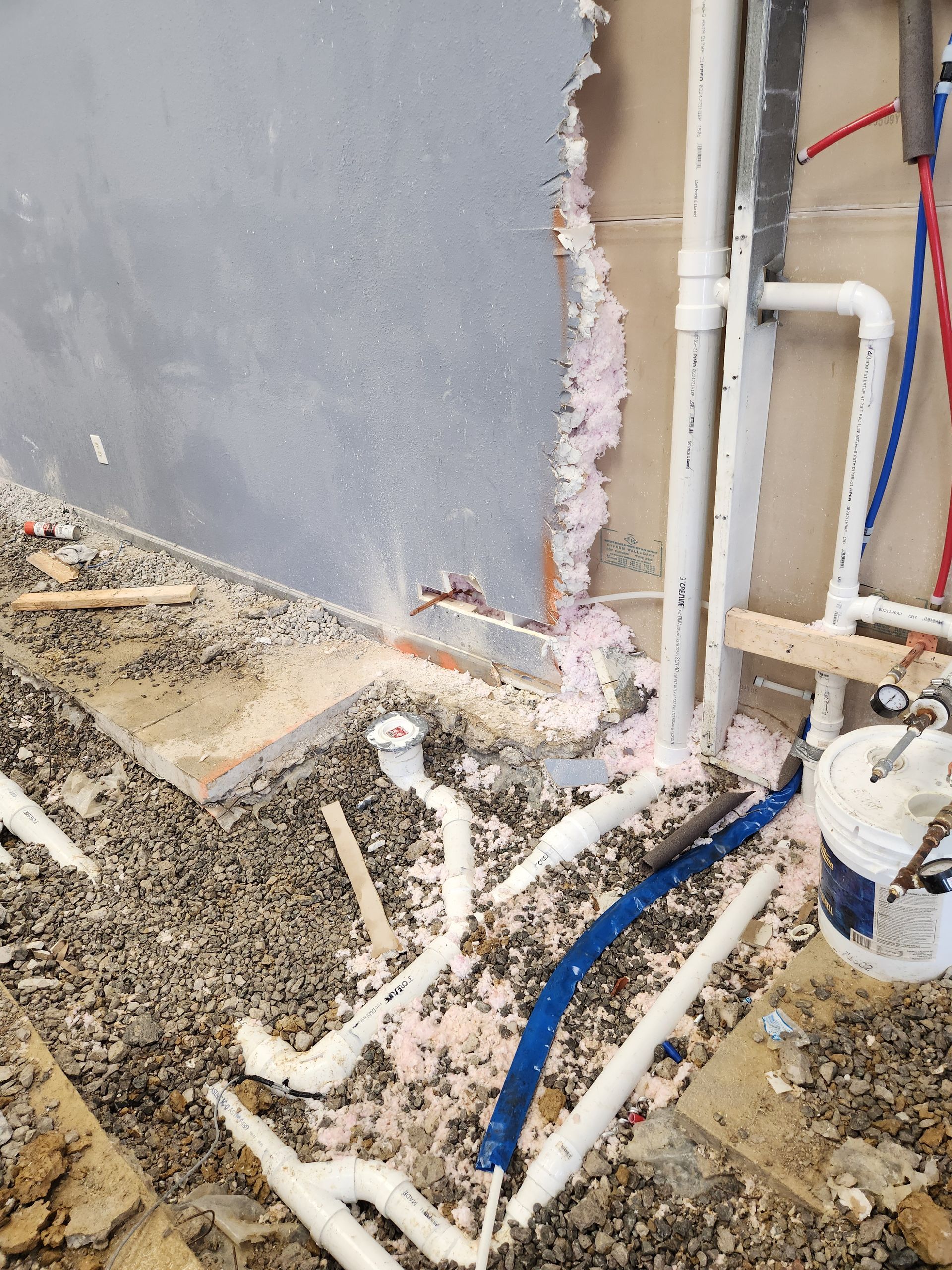 Damaged wall with exposed white and blue pipes in a construction area with debris on the floor