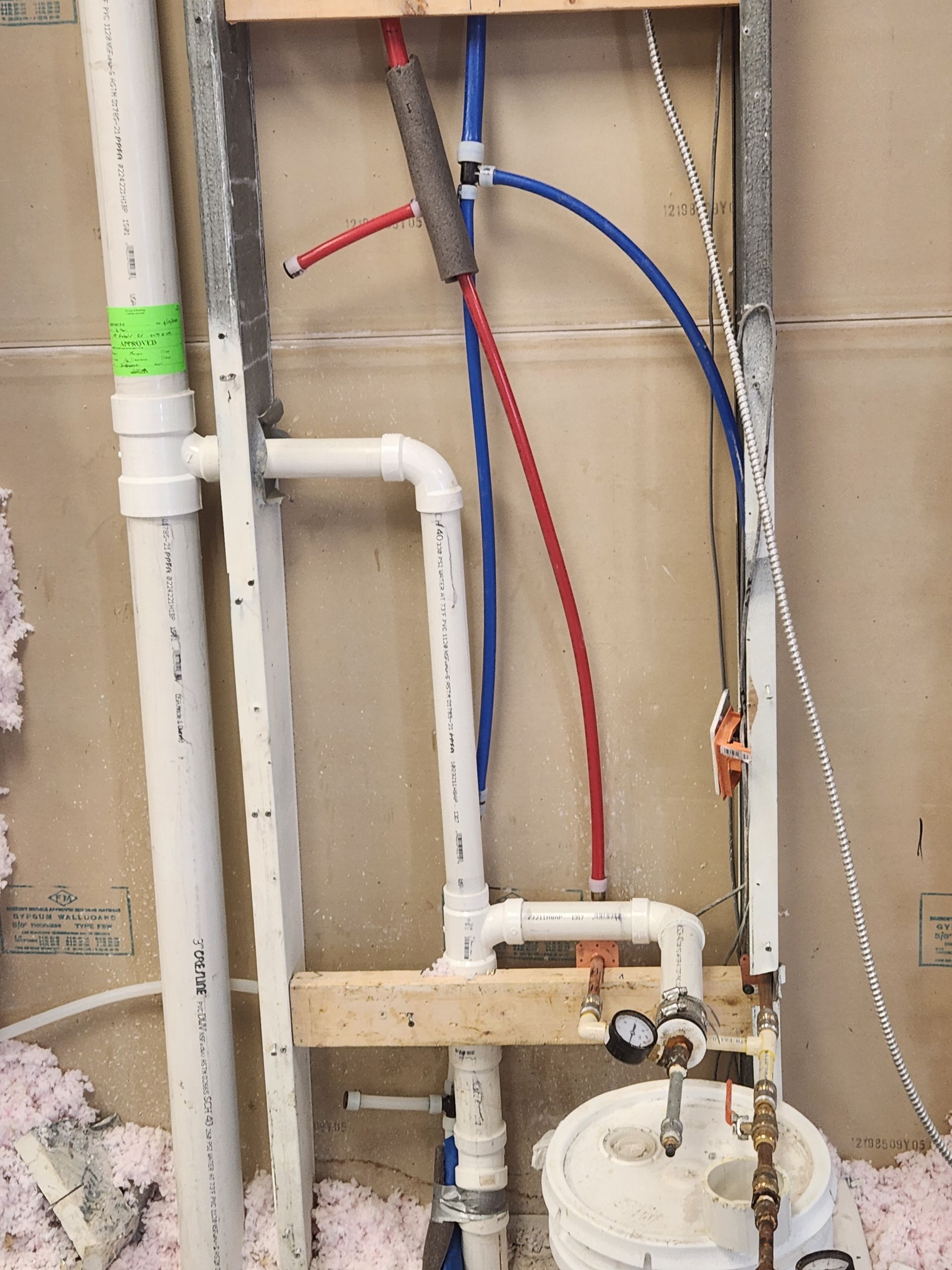 Exposed plumbing in a wall opening with white pipes, red and blue PEX lines, and a water heater below