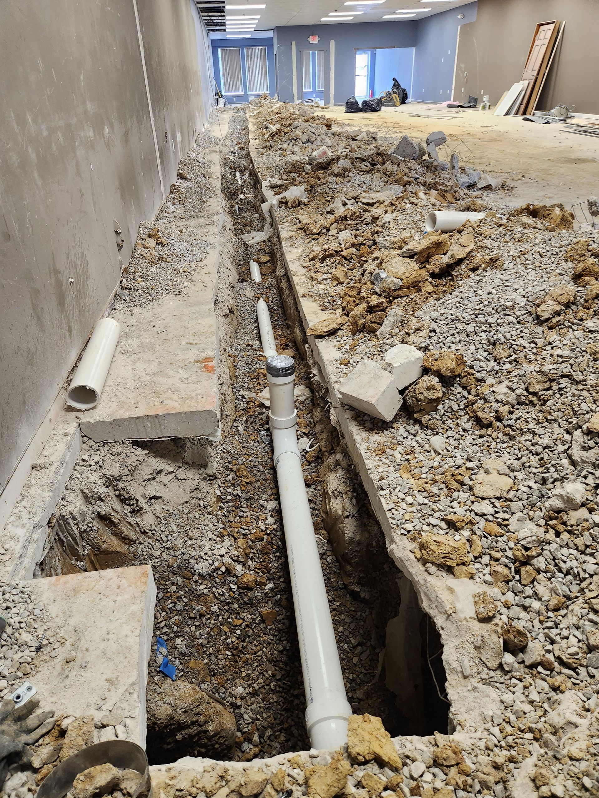 Construction trench with exposed white pipes beside a rough dirt floor and concrete wall in a hallway