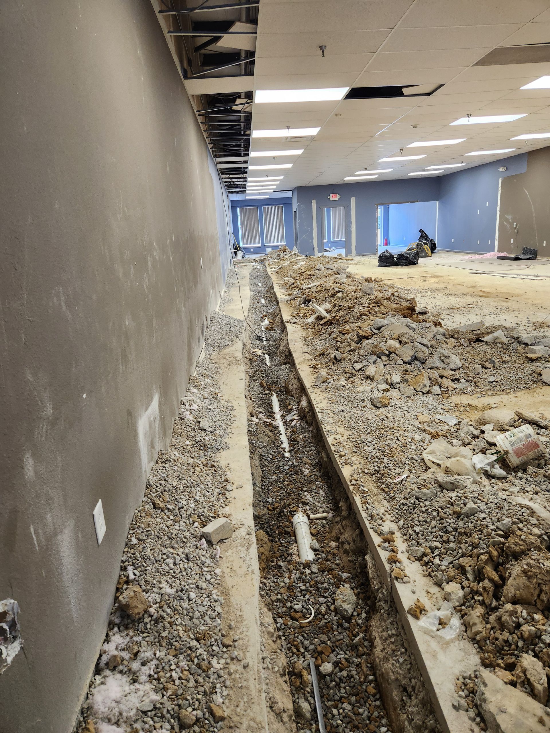 Damaged office interior with exposed floor trench, debris, and unfinished ceiling panels