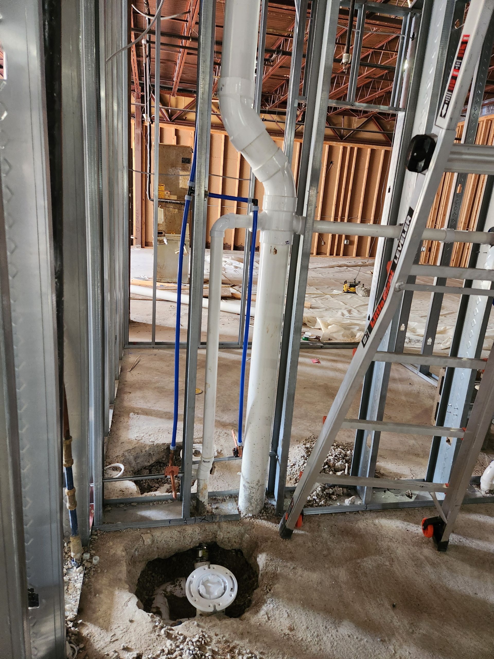 Construction framing with exposed pipes and ladder, showing a floor drain opening in a rough concrete floor.