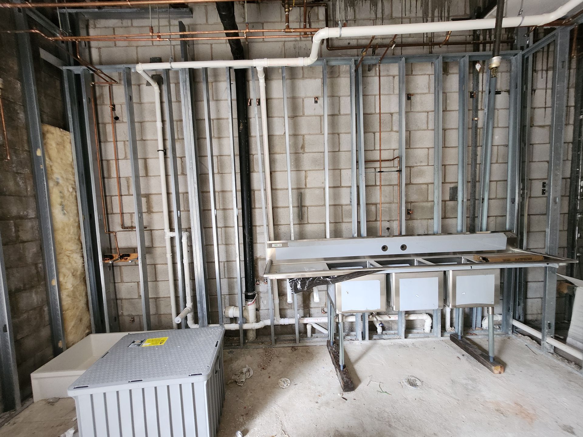 Unfinished utility room with exposed framing, pipes, white sink base, and stacked materials on the floor