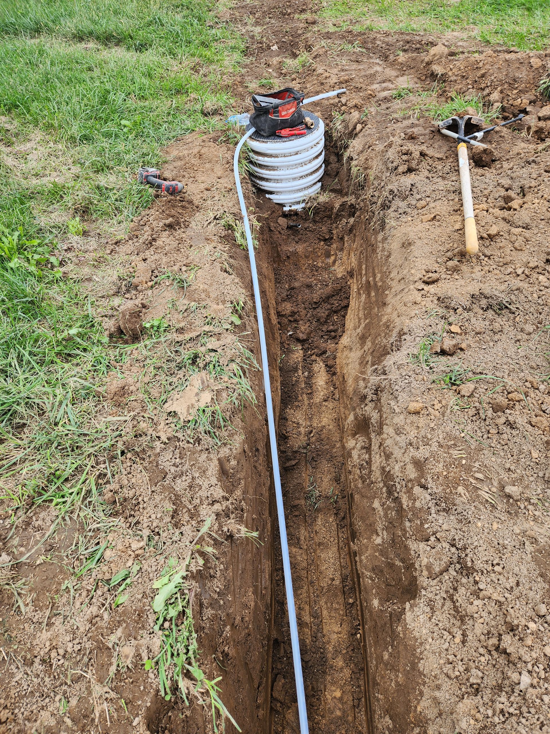 Trenched yard with exposed white pipe and coiled hose beside a shovel on grass and dirt